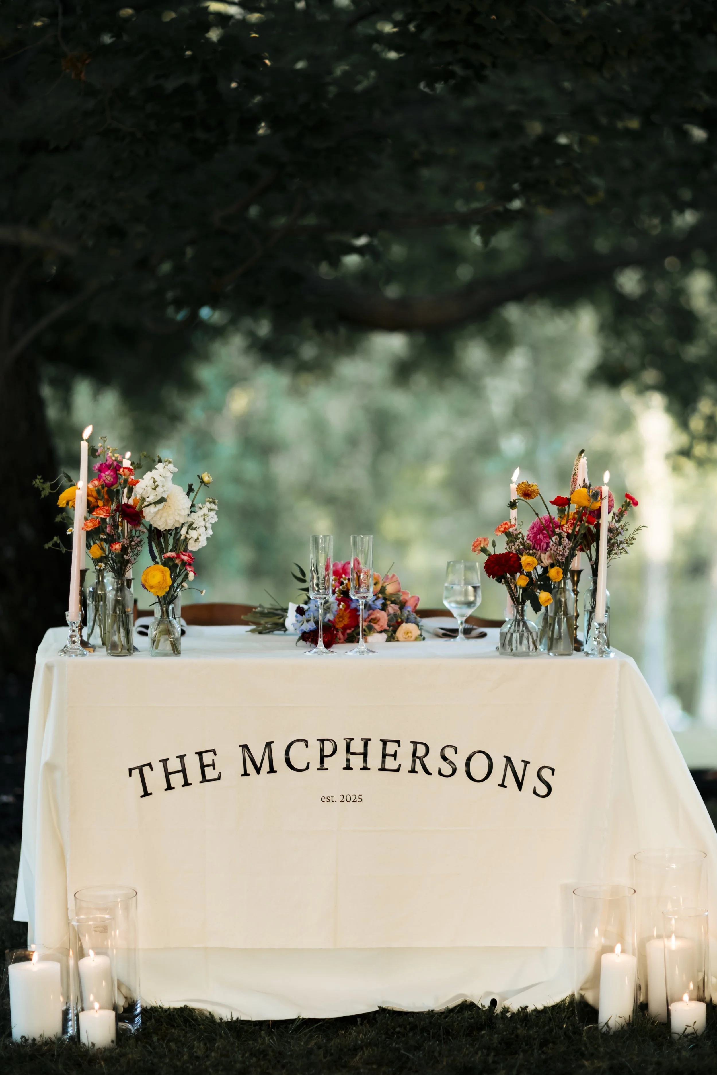 An outdoor wedding reception table with a white tablecloth featuring the names 'The McPhersons' and 'est. 2025'. It is decorated with colorful flower arrangements, tall candles in glass holders, and a glass of water. Candles are placed on the ground 