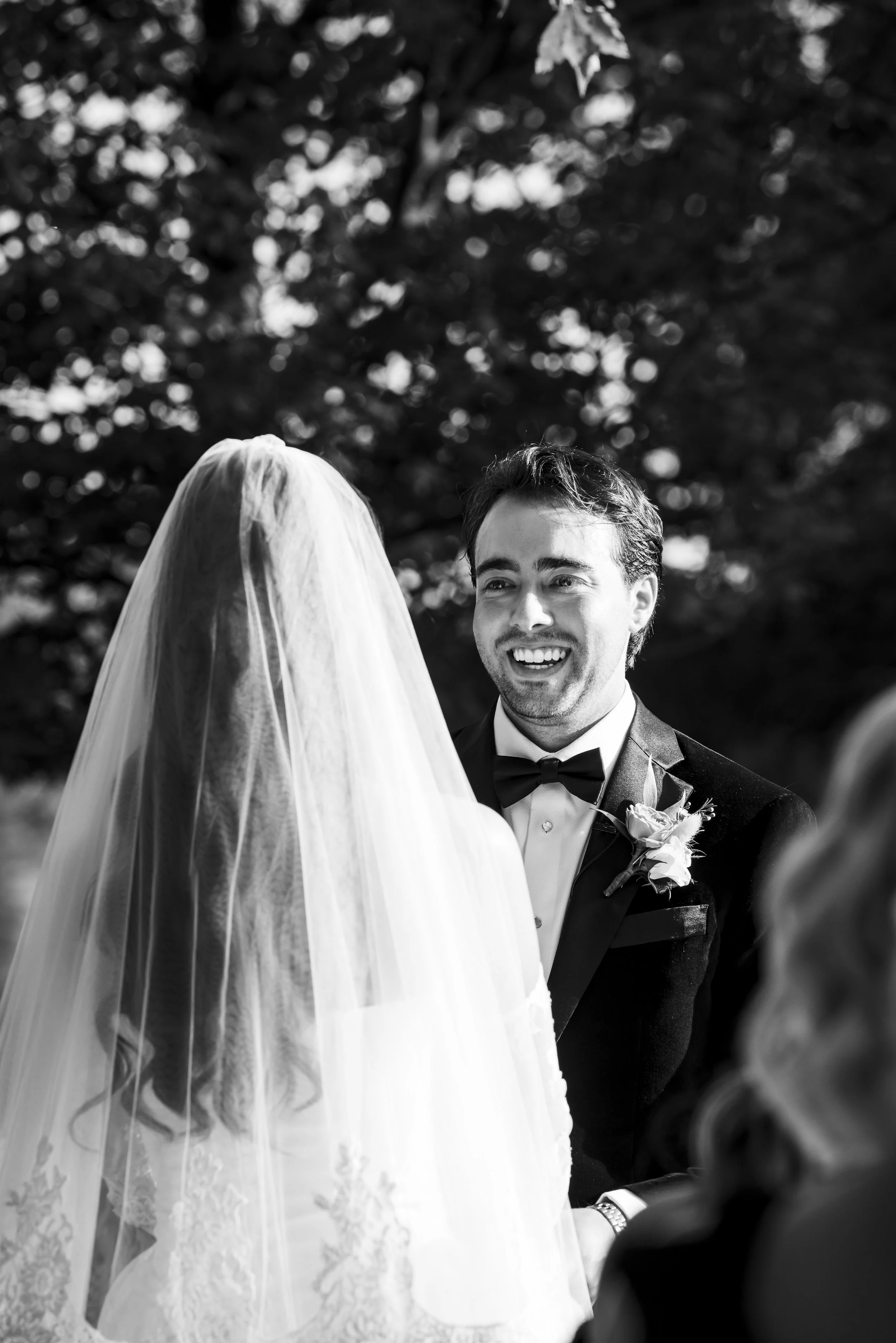 A black and white photograph of a wedding ceremony showing a groom smiling at a bride, who is wearing a veil and lace dress, outdoors with trees in the background.