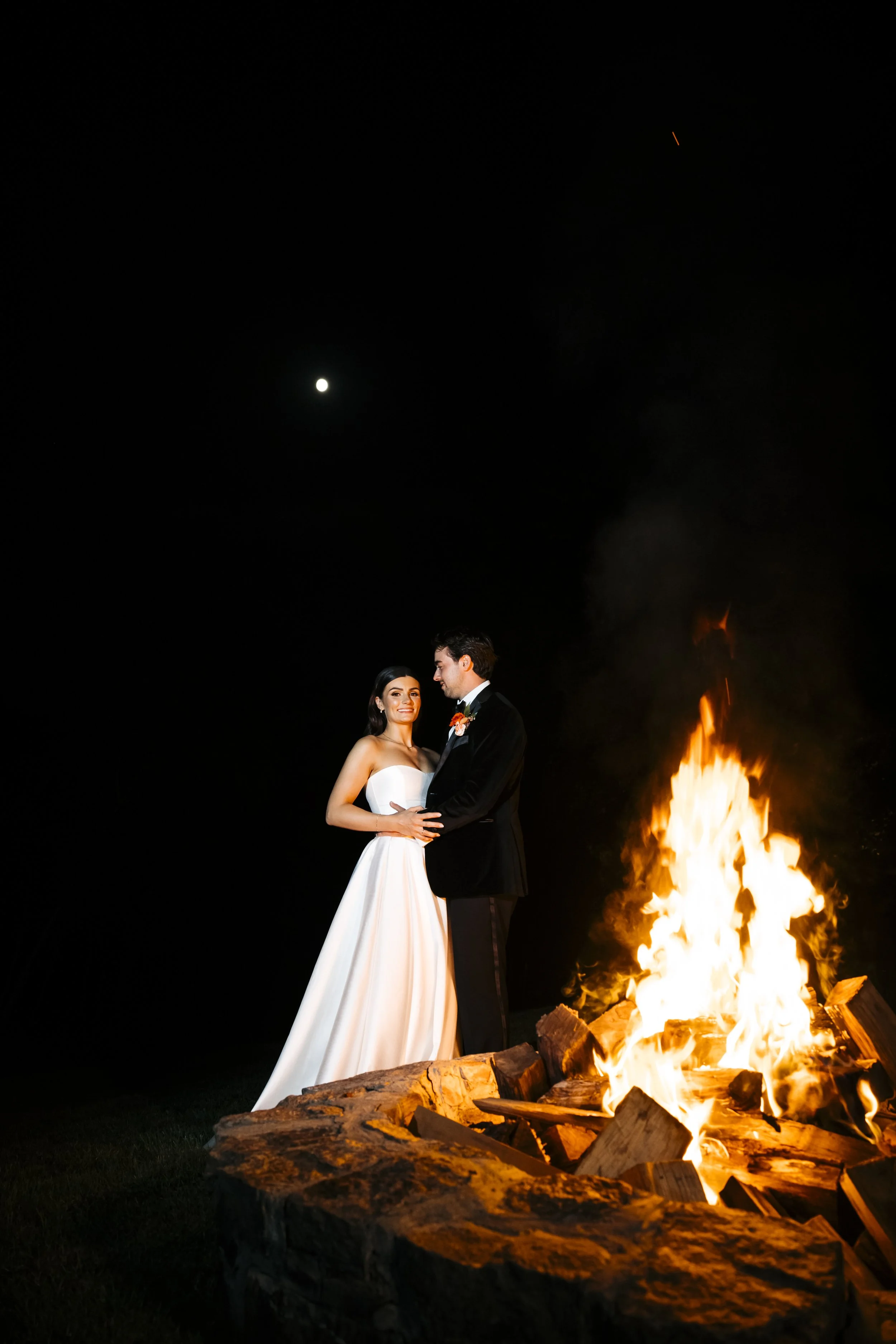 Bride and groom standing close together at night, holding hands, in front of a large bonfire on a stone fire pit, with a full moon in the dark sky.