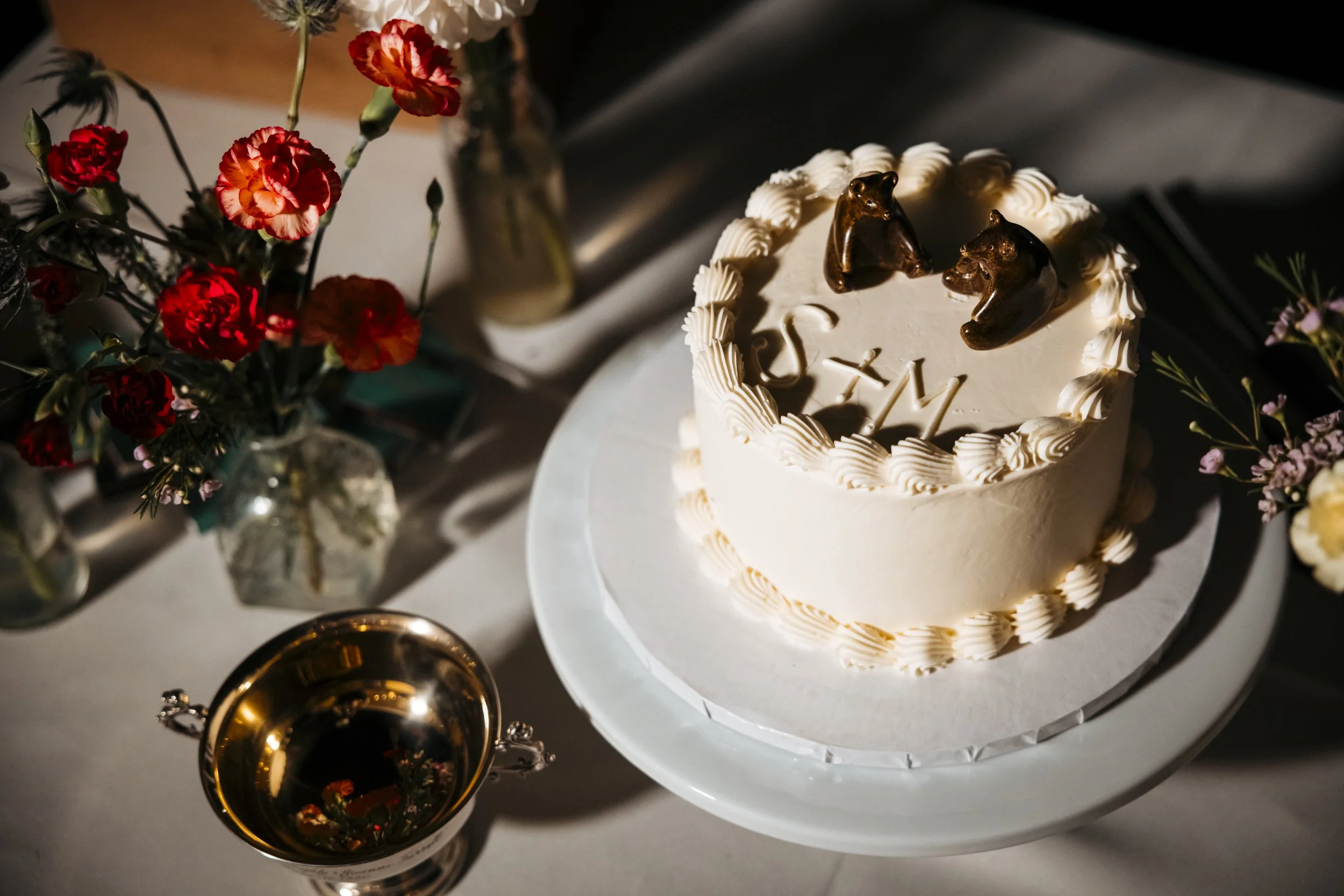 A white frosted cake decorated with chocolate horse head toppers and cream piped border, surrounded by flower arrangements in small vases on a table.