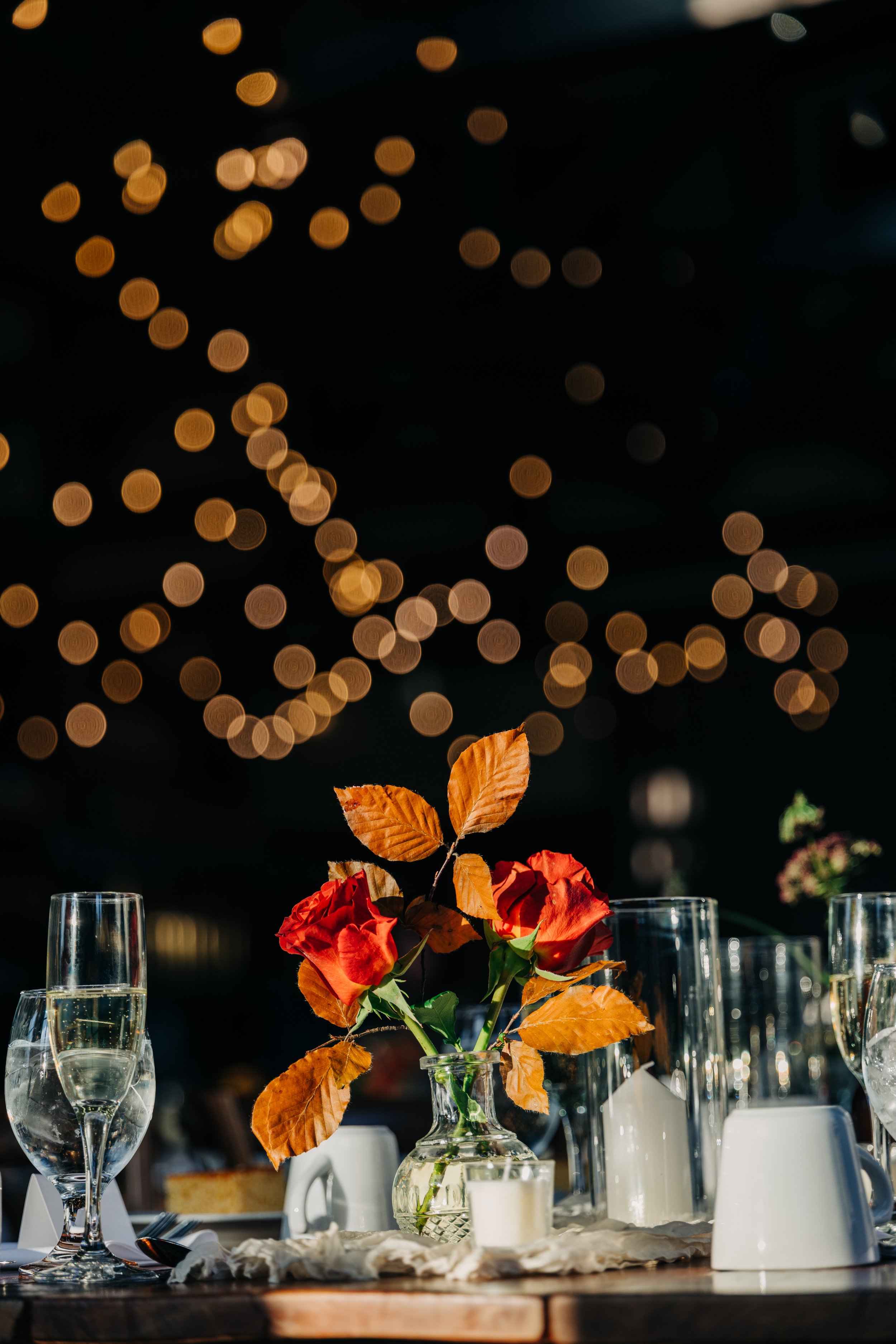 A dining table centerpiece with red roses and orange leaves in a glass vase, surrounded by champagne glasses, white candles, and white cups, with string lights in the background creating a bokeh effect.