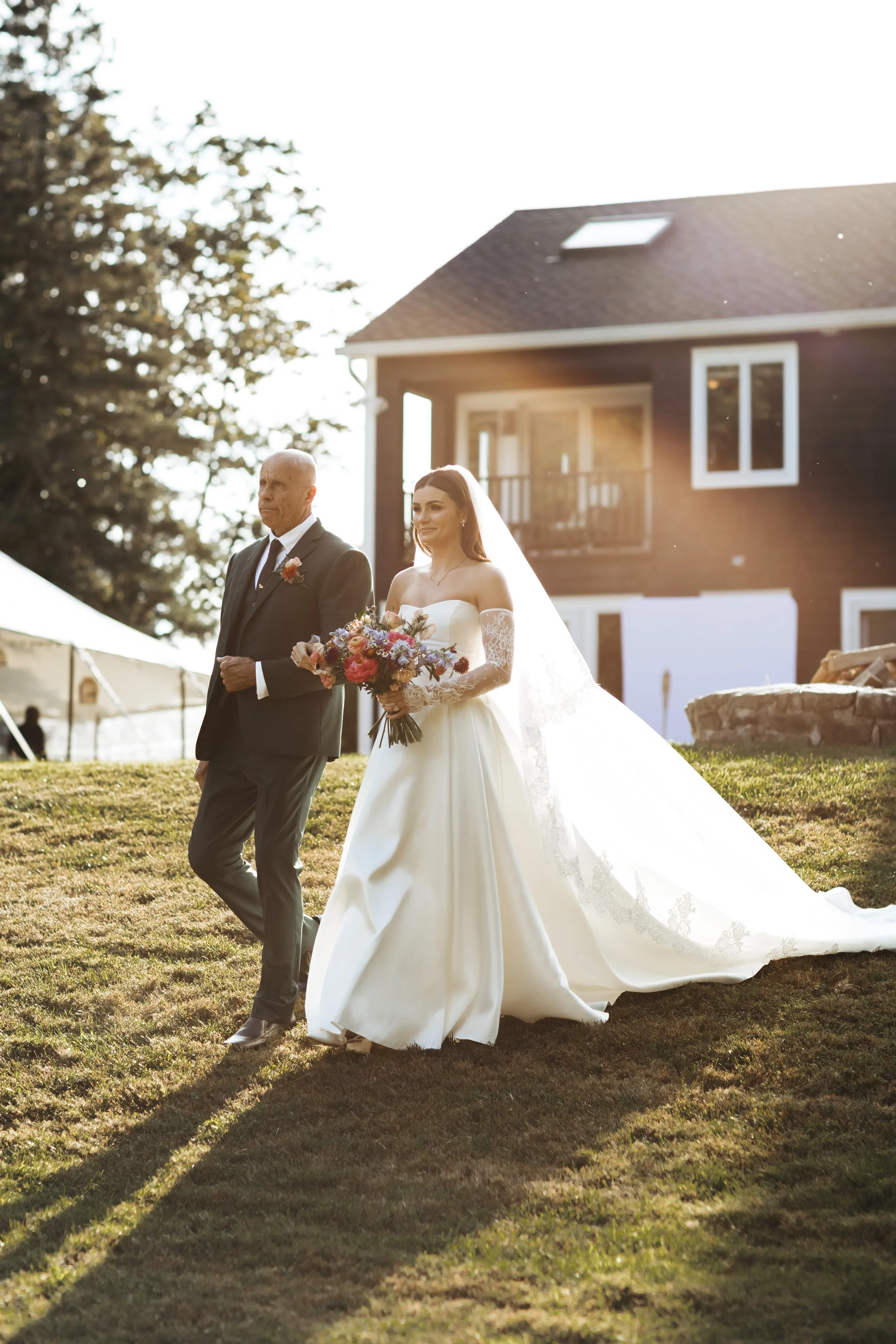 Bride walking outside with her father, holding a bouquet, during a wedding ceremony at sunset.