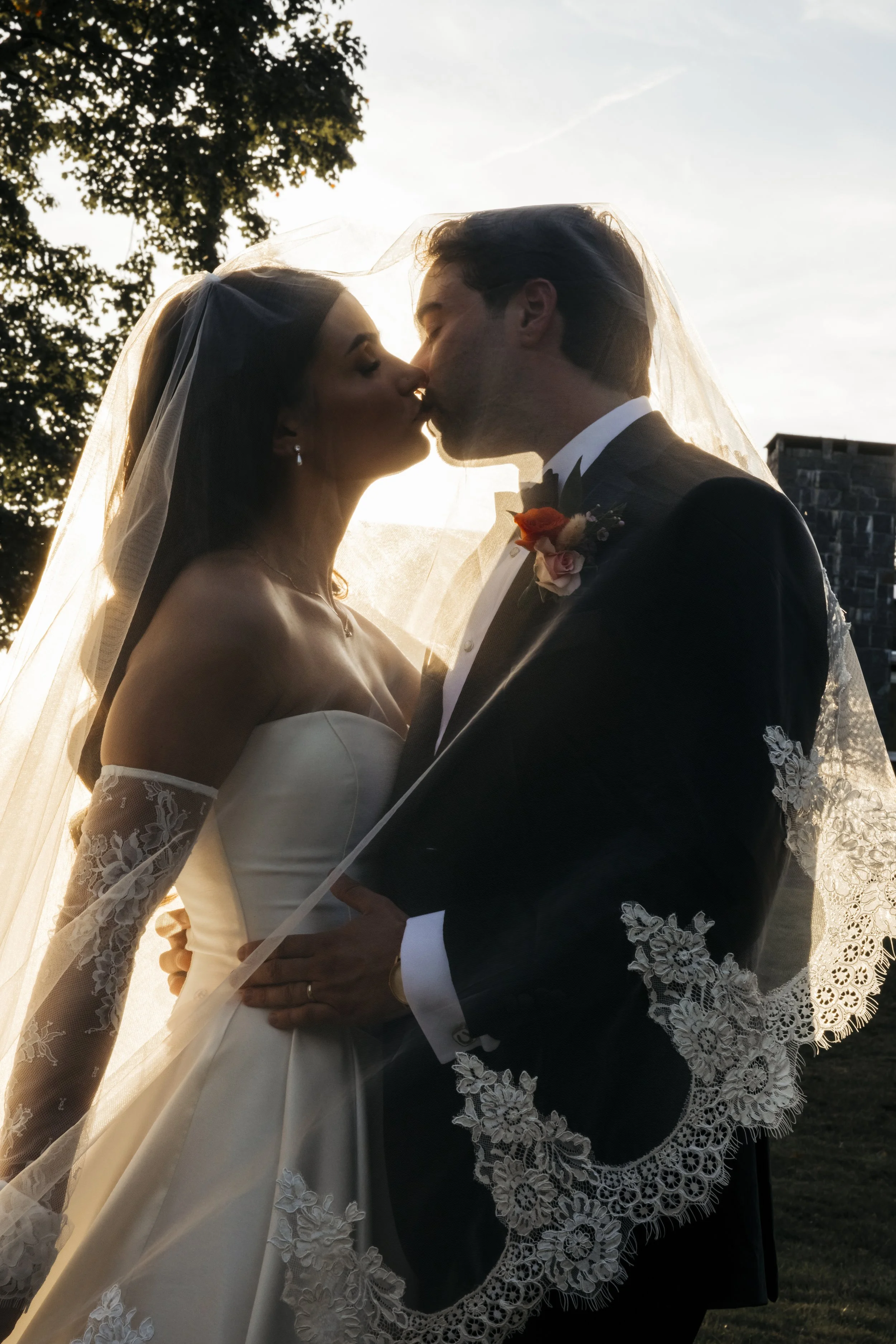 A bride and groom in wedding attire sharing a kiss under a veil during sunset outdoors.