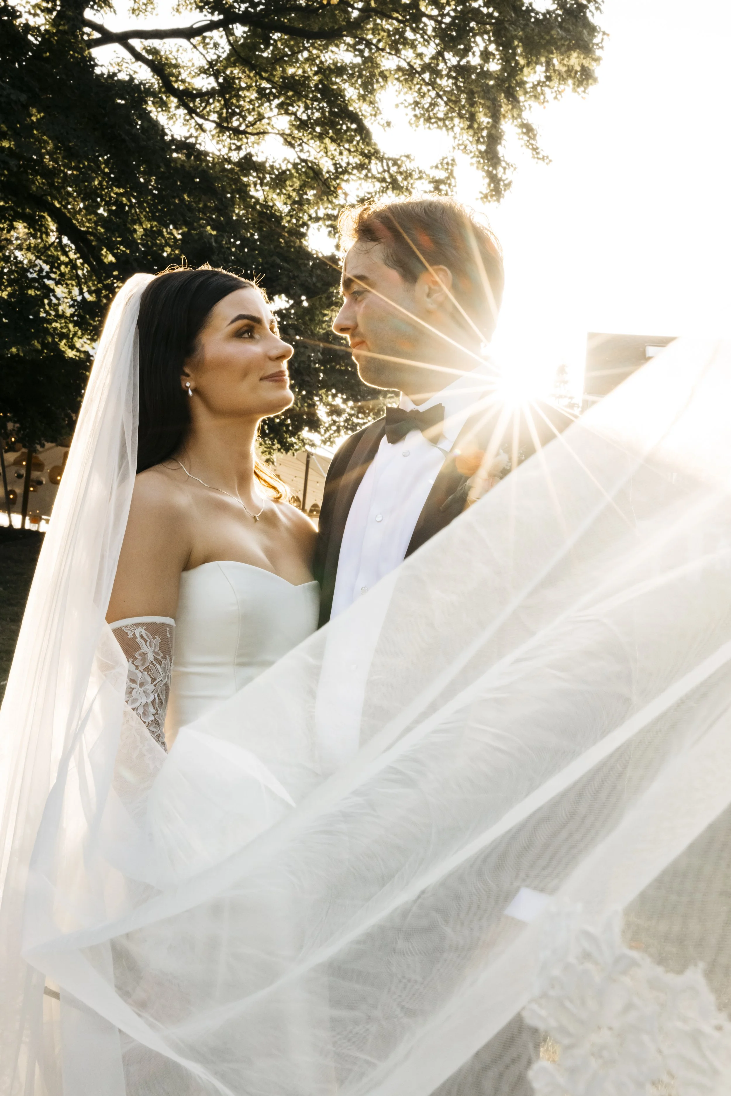 Bride and groom standing outdoors during sunset, gazing at each other, with sunlight creating a starburst effect behind them.