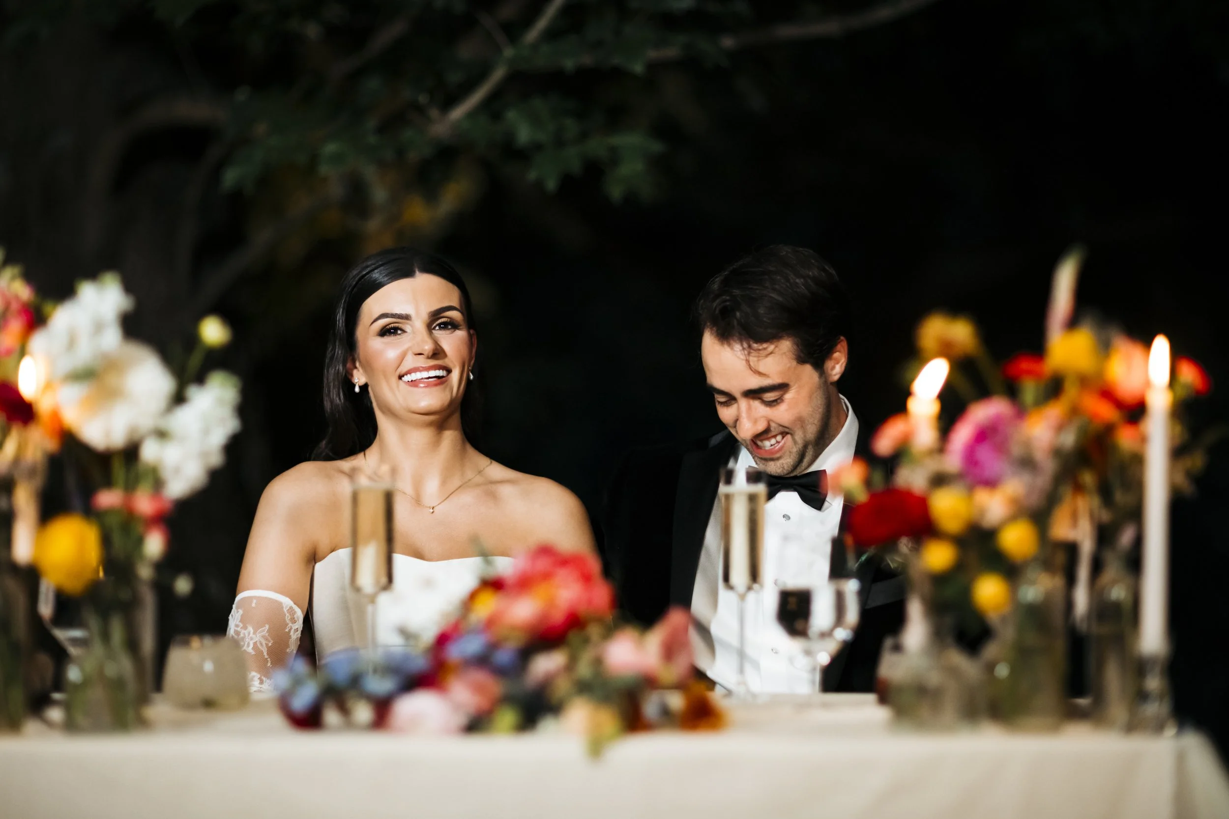 A bride and groom sitting at a wedding reception table, smiling and looking joyful. The bride has long dark hair and is wearing a strapless dress with lace sleeves, while the groom has dark hair and is wearing a black tuxedo with a bow tie. The table