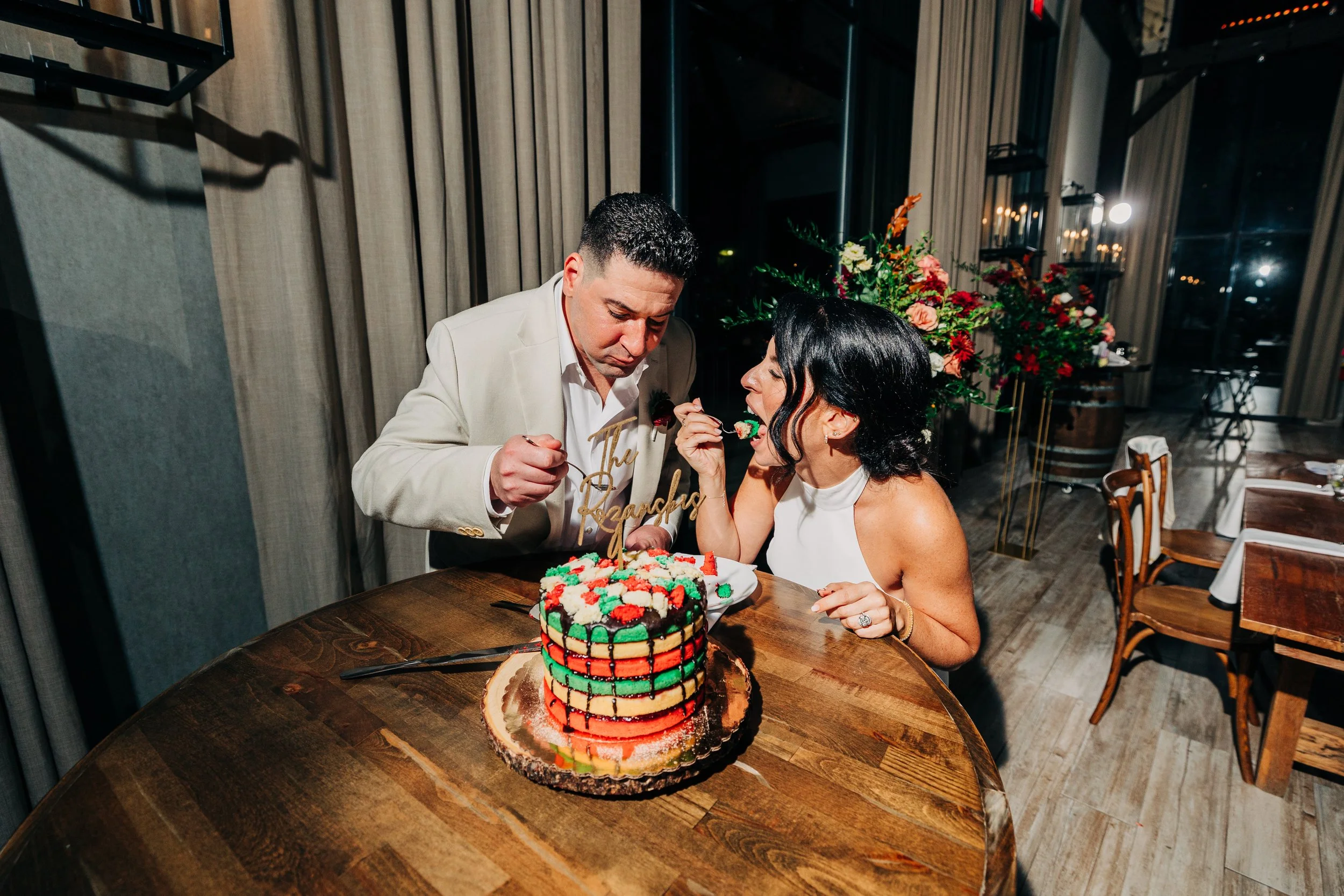 A couple dressed in elegant attire celebrating a birthday with a colorful layered cake on a wooden table, the woman feeding the man a piece of cake, and a floral arrangement in the background.