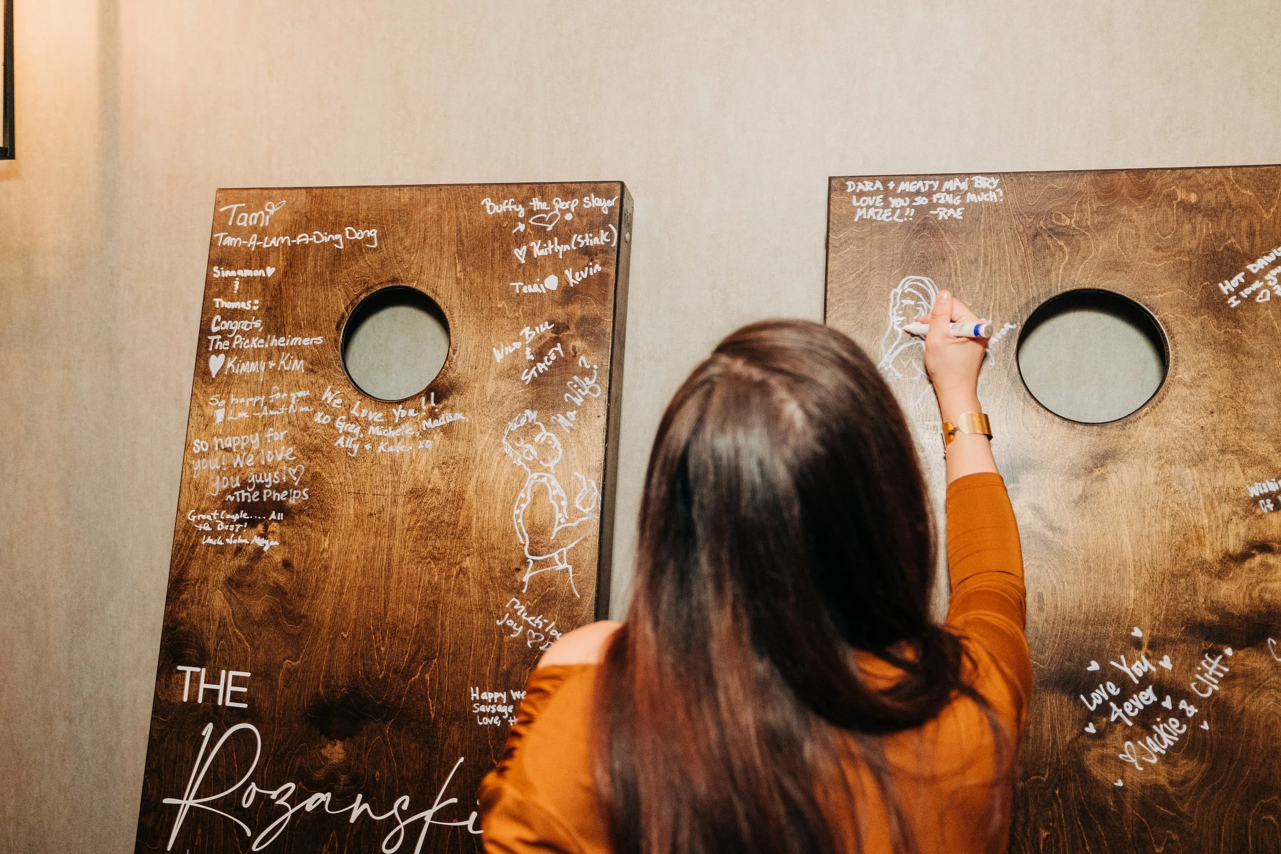 A woman with long brown hair, wearing an orange top, writes on wooden boards with white markers. The boards have handwritten messages, drawings, and signatures, with circular mirrors in the centers.