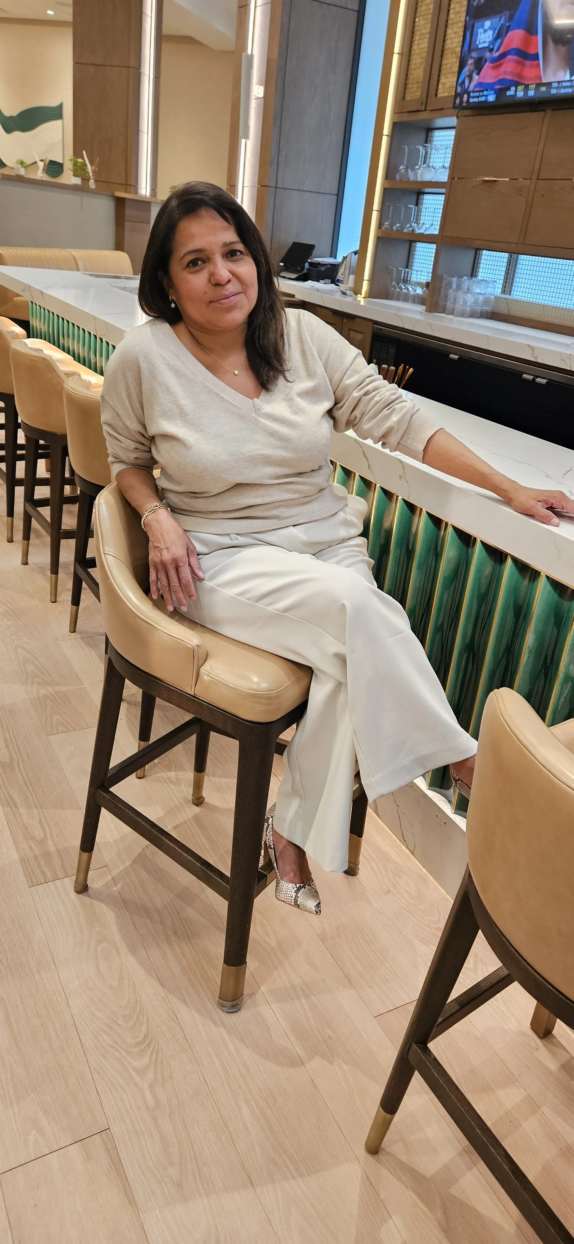 A woman with shoulder-length dark hair sitting on a bar stool in a restaurant or bar. She is wearing a beige sweater, white pants, and high-heeled shoes. The background includes a marble bar counter, glass shelves with glasses, a television, and wooden decor.