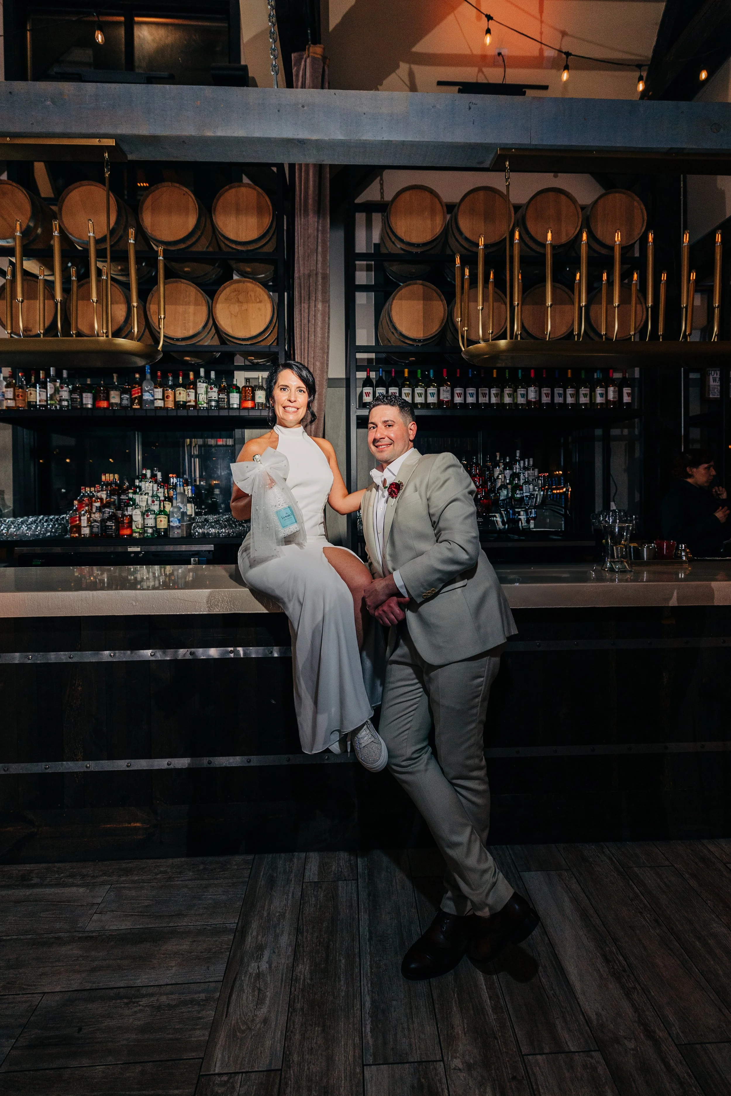 A woman in a white dress sitting on a bar counter and a man in a beige suit standing next to her, smiling at the camera in a bar or restaurant setting with alcohol bottles and barrels in the background.