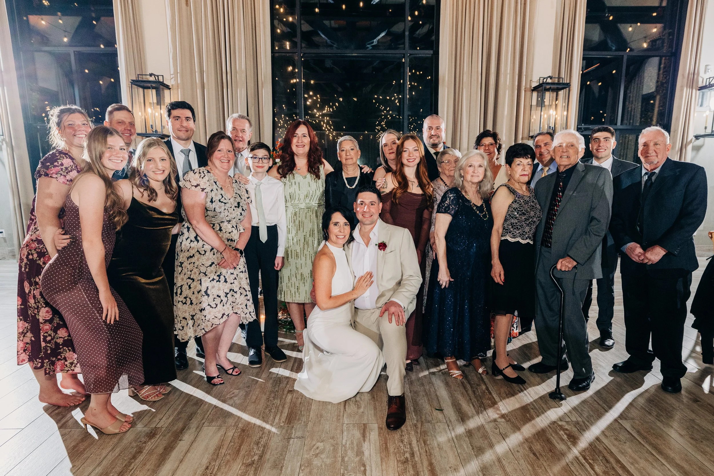 Group of people at a wedding celebration, including a bride and groom in wedding attire, standing and smiling indoors with a large window and curtains in the background.