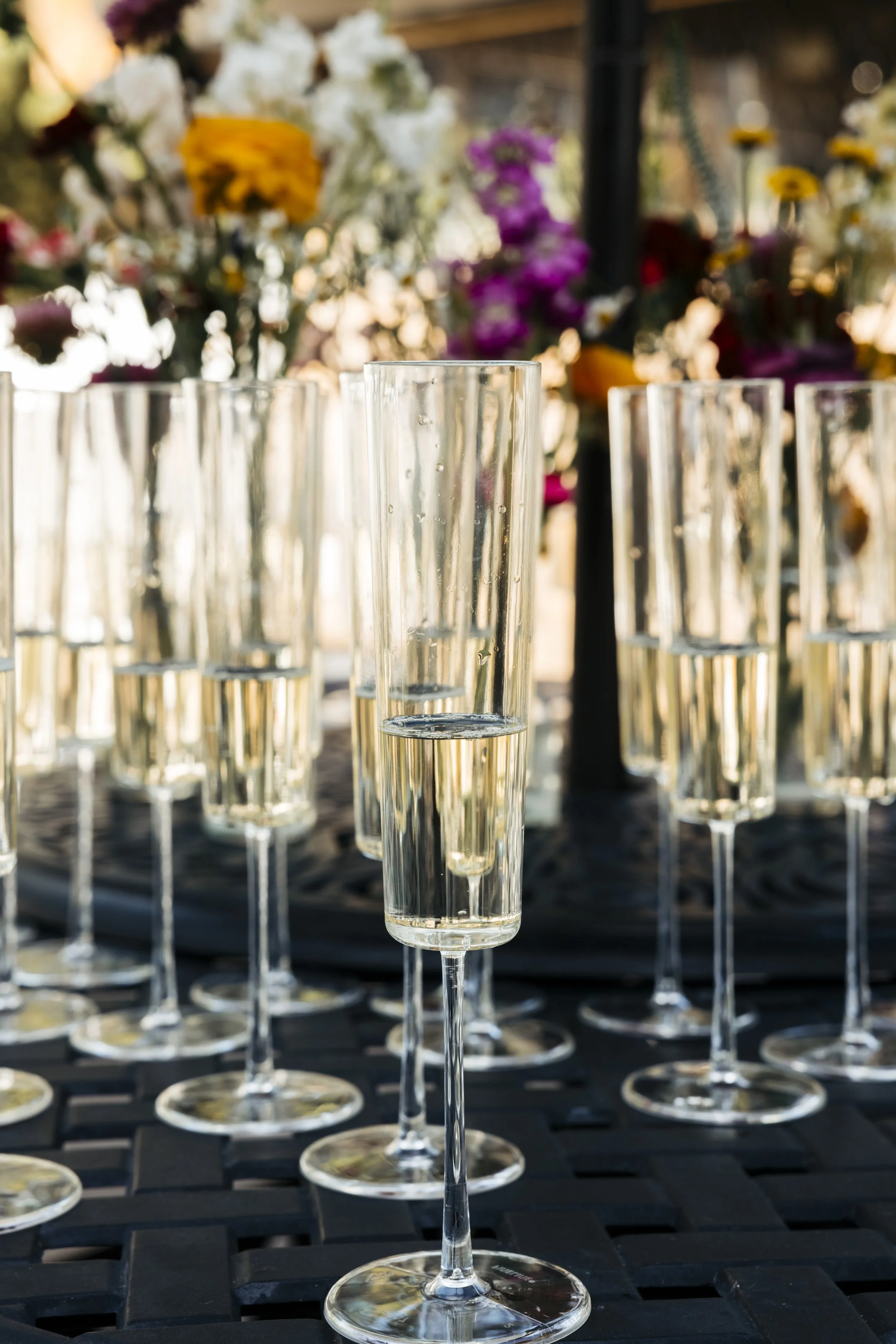Multiple champagne flutes with bubbly, set on a black surface, with colorful flowers in the background.