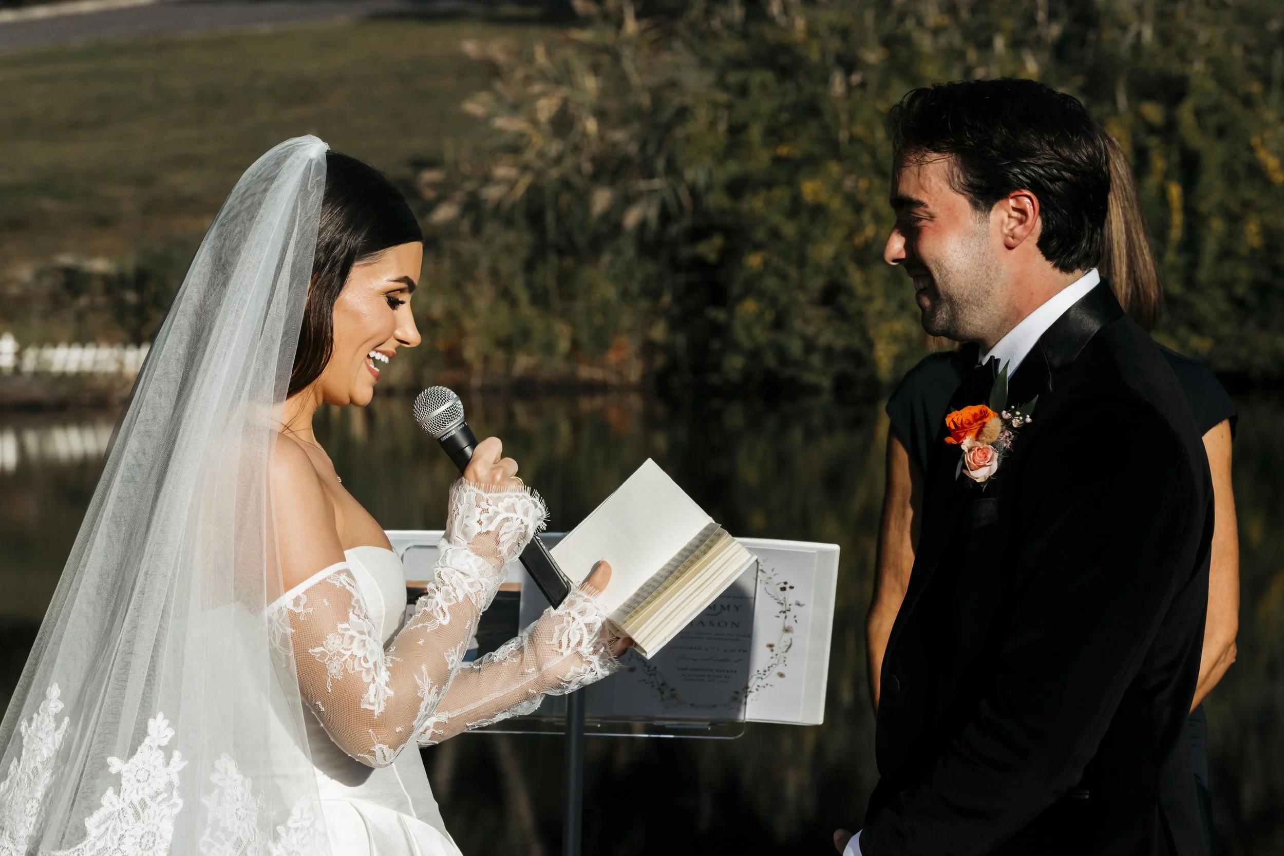A bride and groom exchanging vows outdoors near a body of water during a wedding ceremony, with the bride reading her vows from a notebook and holding a microphone.