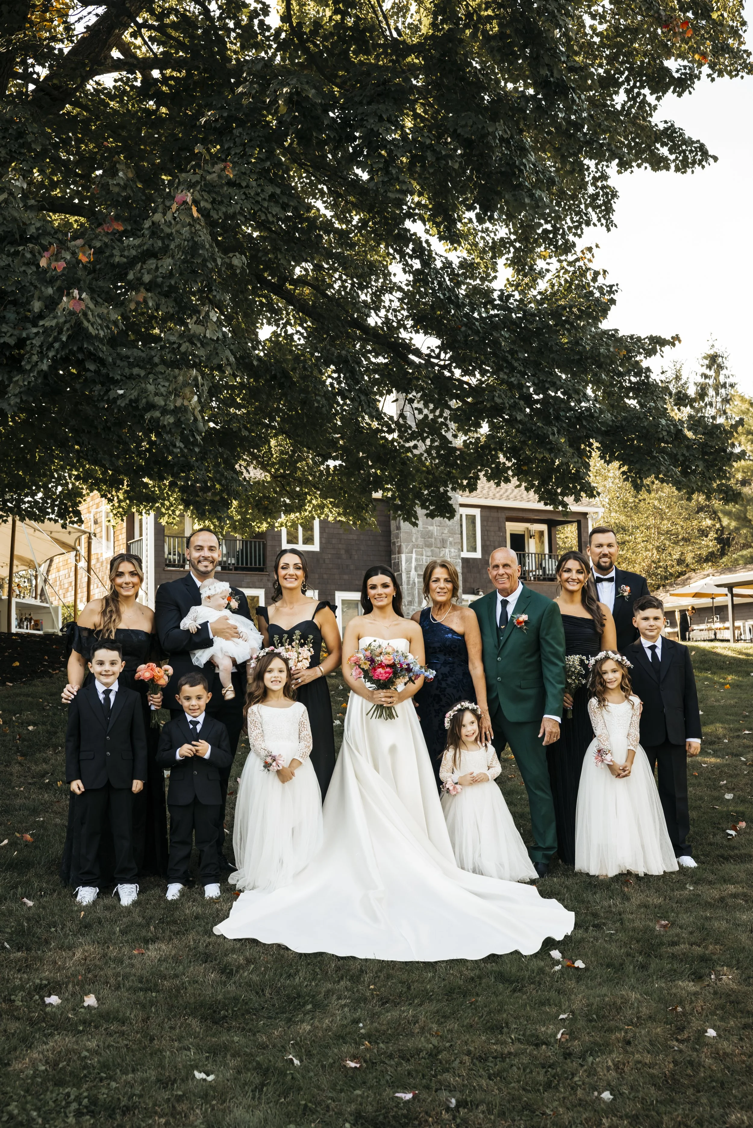 A group of people dressed in formal wedding attire, including a bride and groom, standing outdoors in front of a house with trees and a grassy yard.