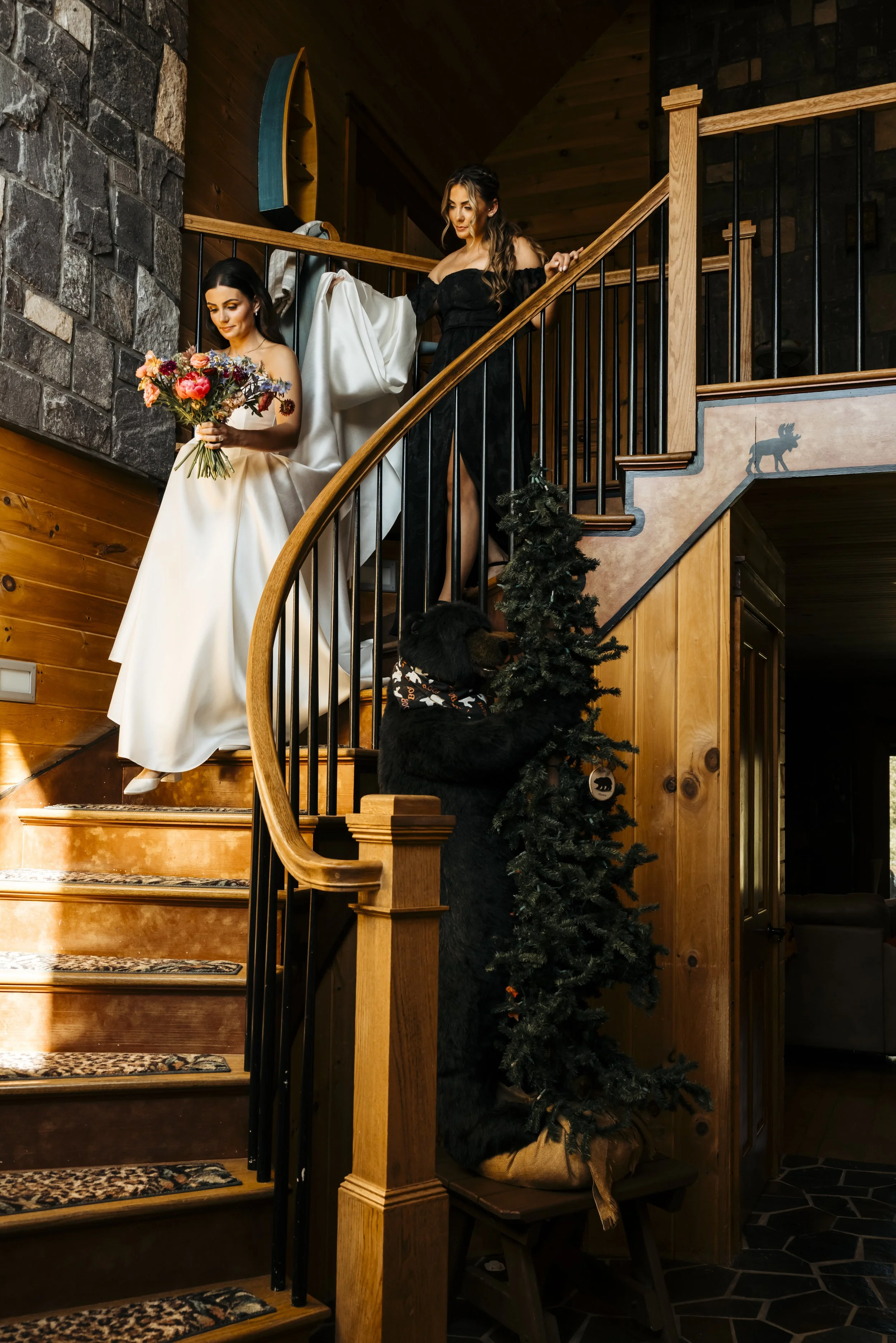 Two women on a wooden staircase, with one holding a bouquet of flowers and the other holding a white cloth, and a black dog at the bottom of the stairs.