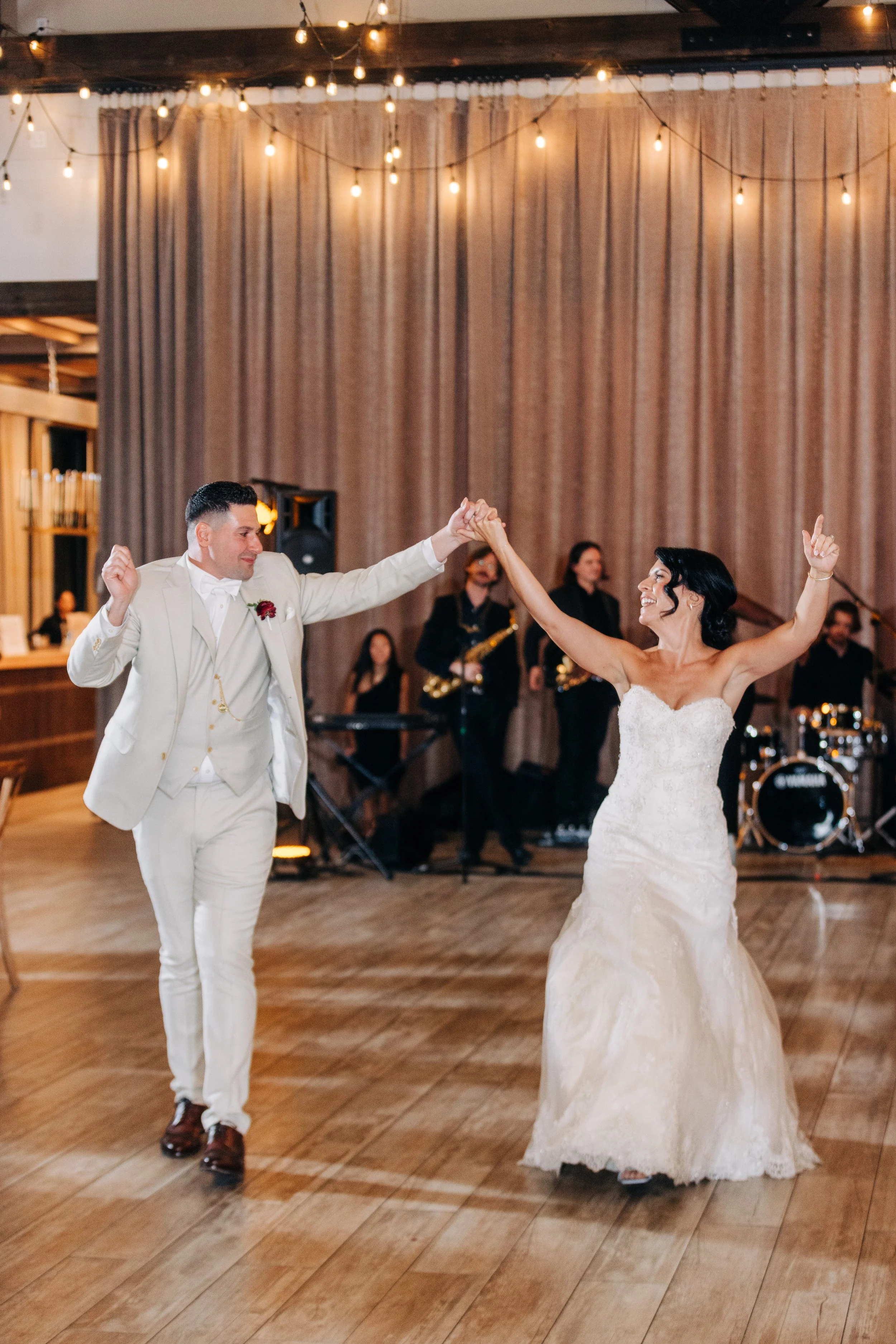 A bride and groom dancing at their wedding reception. The groom is dressed in a white tuxedo with a boutonniere, and the bride is in a strapless white wedding gown. They are holding hands and smiling, with the bride raising her other arm. There are live musicians in the background playing instruments, and the venue has warm lighting with string lights and beige curtains.