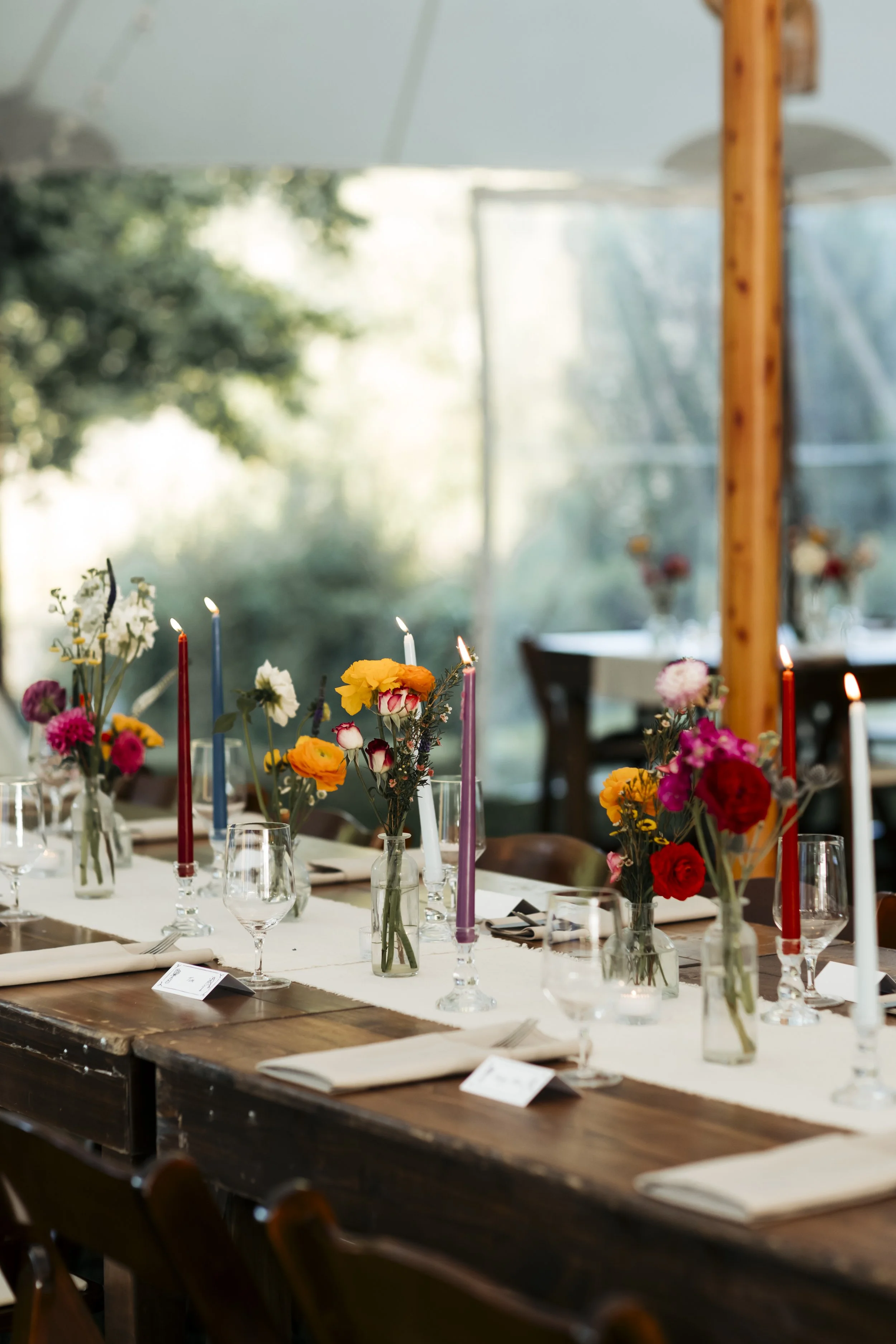 A dining table decorated with colorful flower arrangements in glass vases and tall, lit candles, set with napkins, wine glasses, and place cards in a well-lit, outdoor or greenhouse setting.