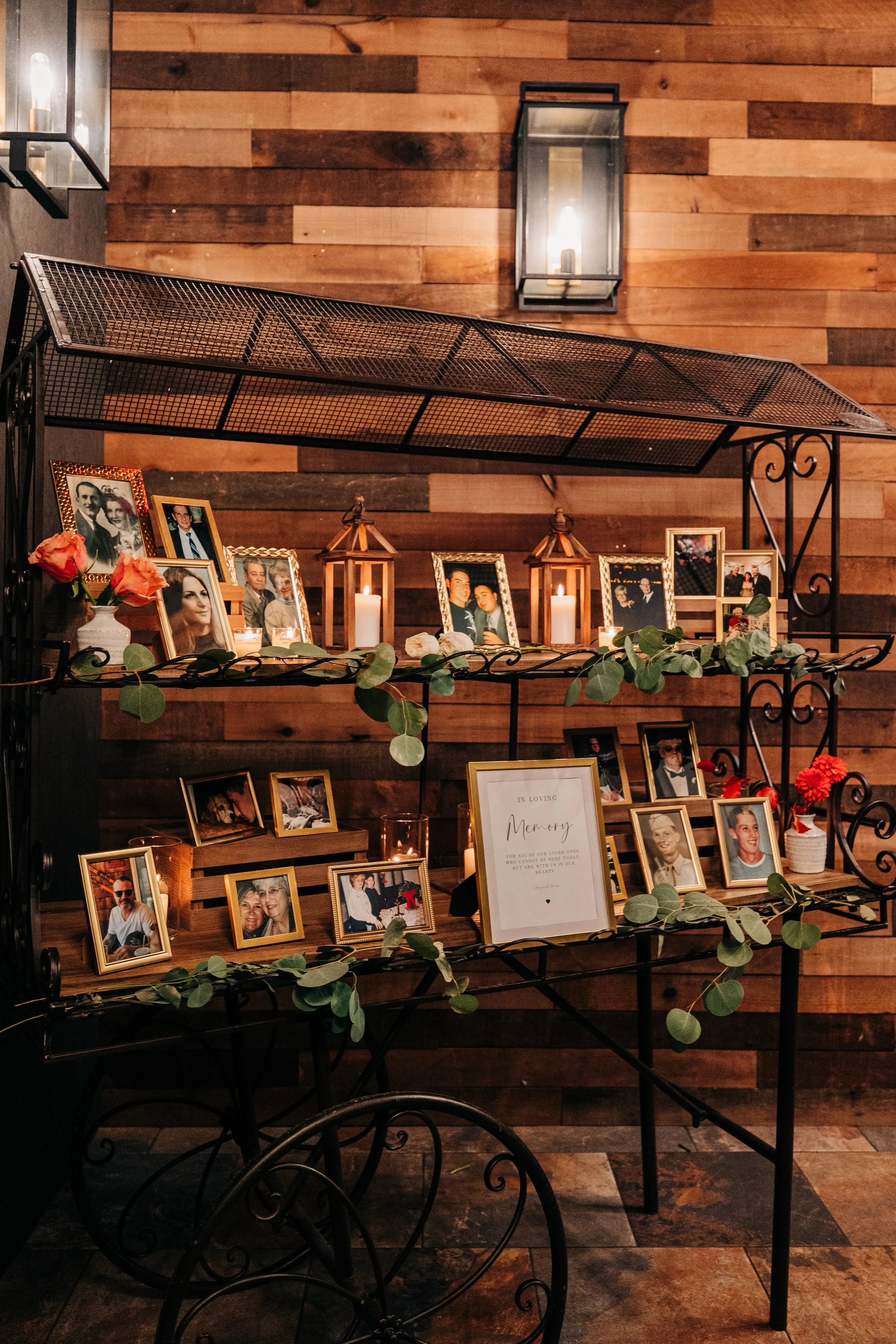 Memorial display with framed photographs of loved ones, lit candles, and decorative greenery on black metal shelves against a wooden wall.