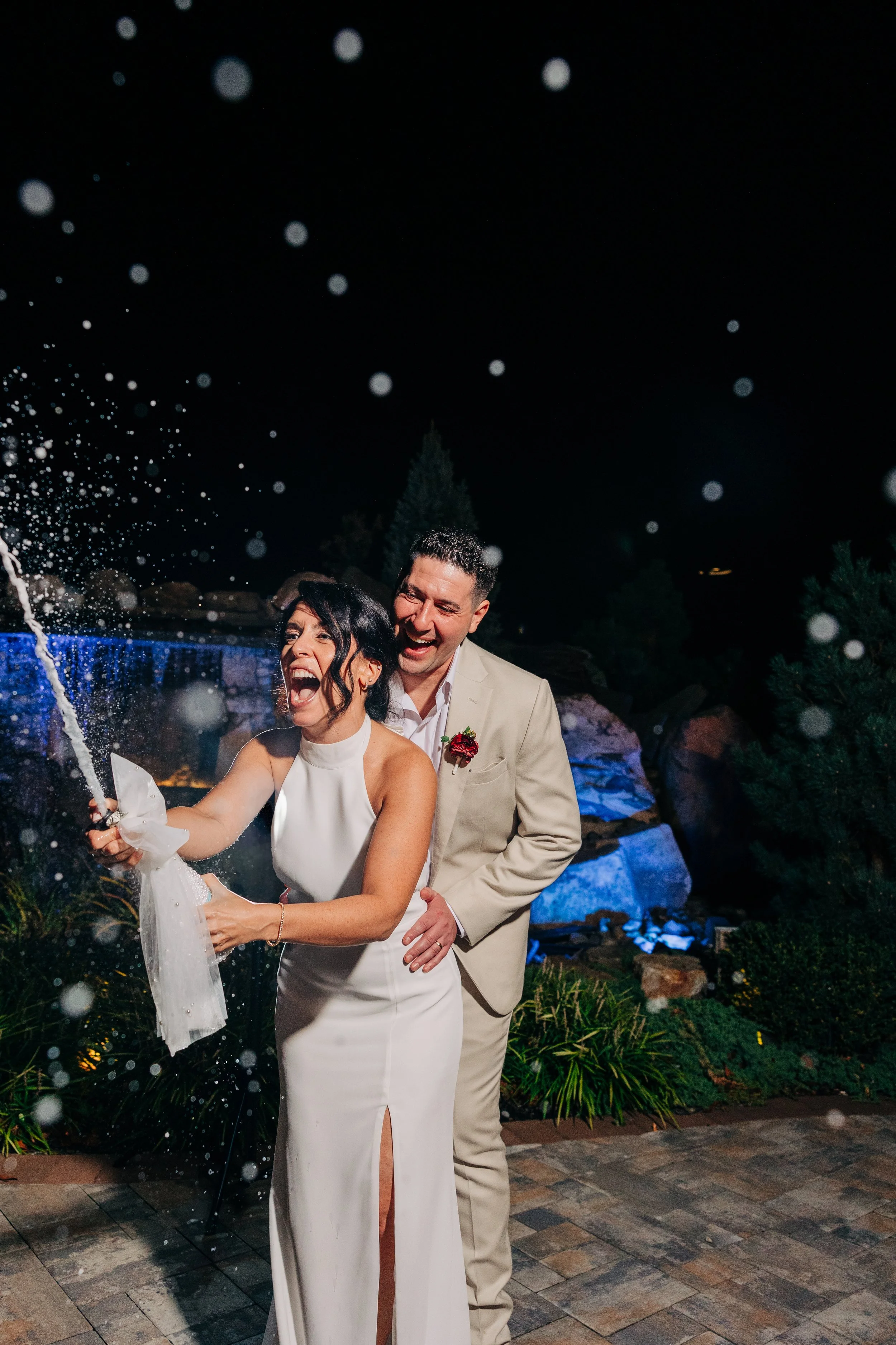 A newlywed couple celebrating, with the bride opening a champagne bottle, surrounded by bubbles, at night outdoor setting.