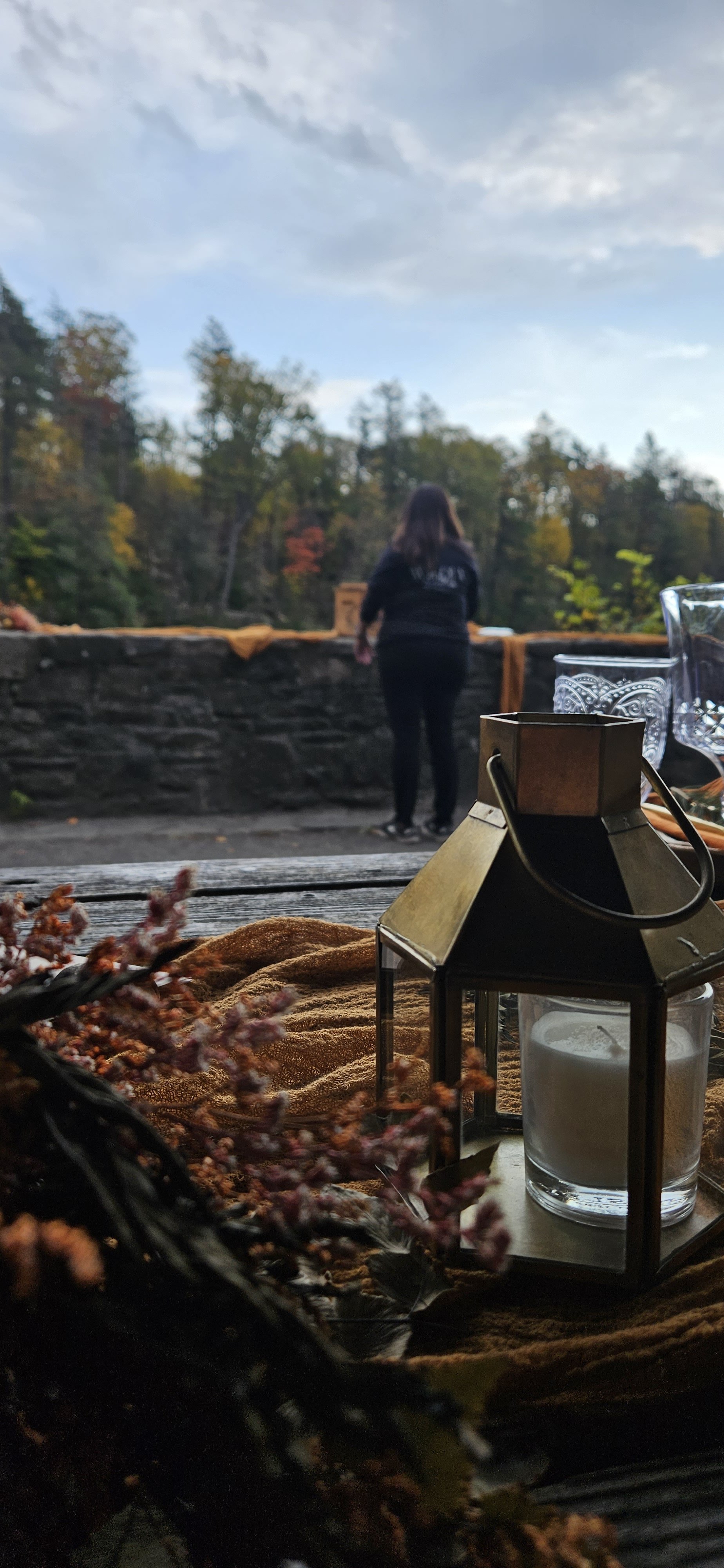 A table with a candle in a glass holder and decorative glassware, with a lantern, and fall-themed decorations, looking out onto a woman standing outdoors in front of a stone wall with trees in the background.