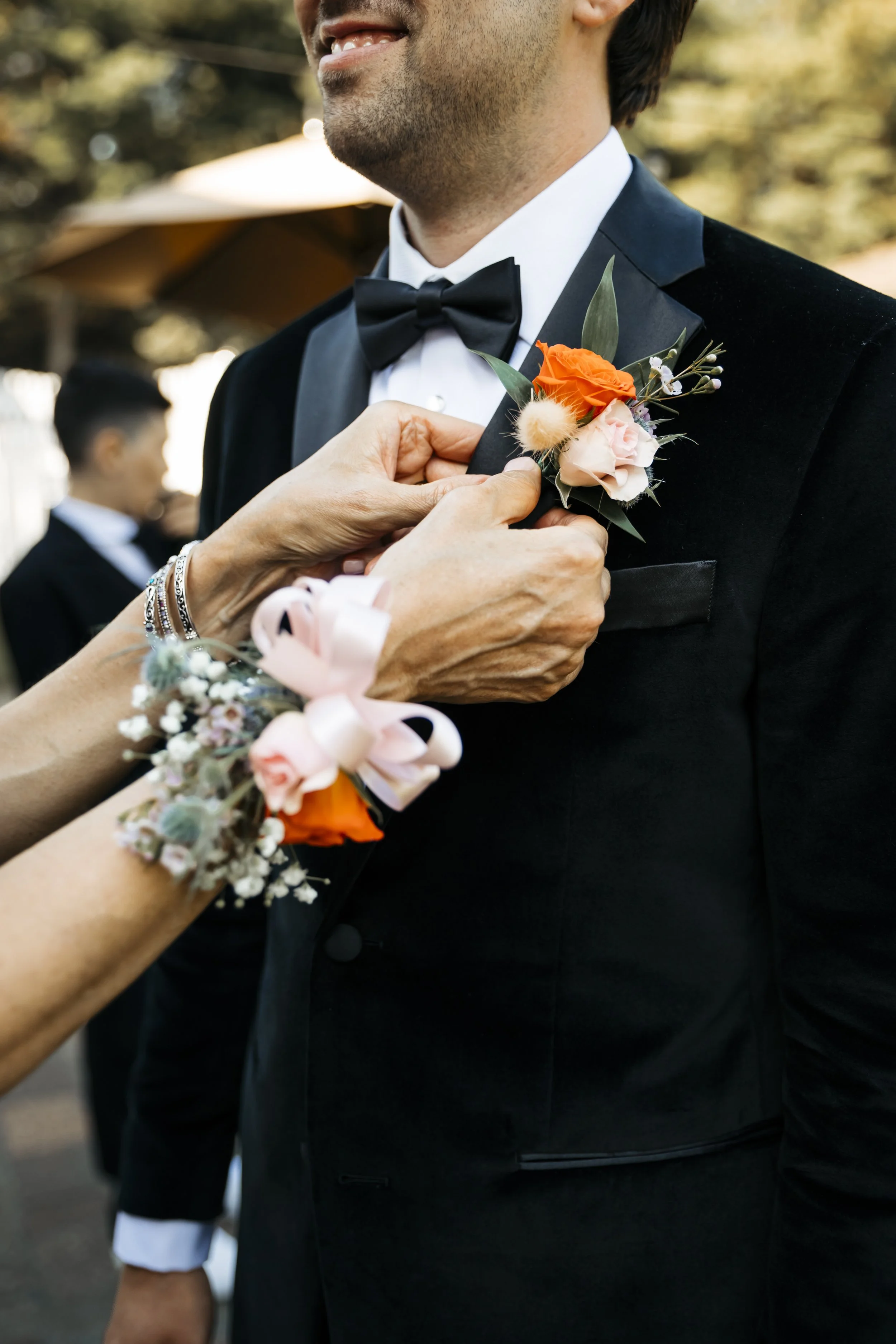 Person in a black tuxedo with a bow tie has a small boutonniere being pinned on their lapel during a wedding or formal event.