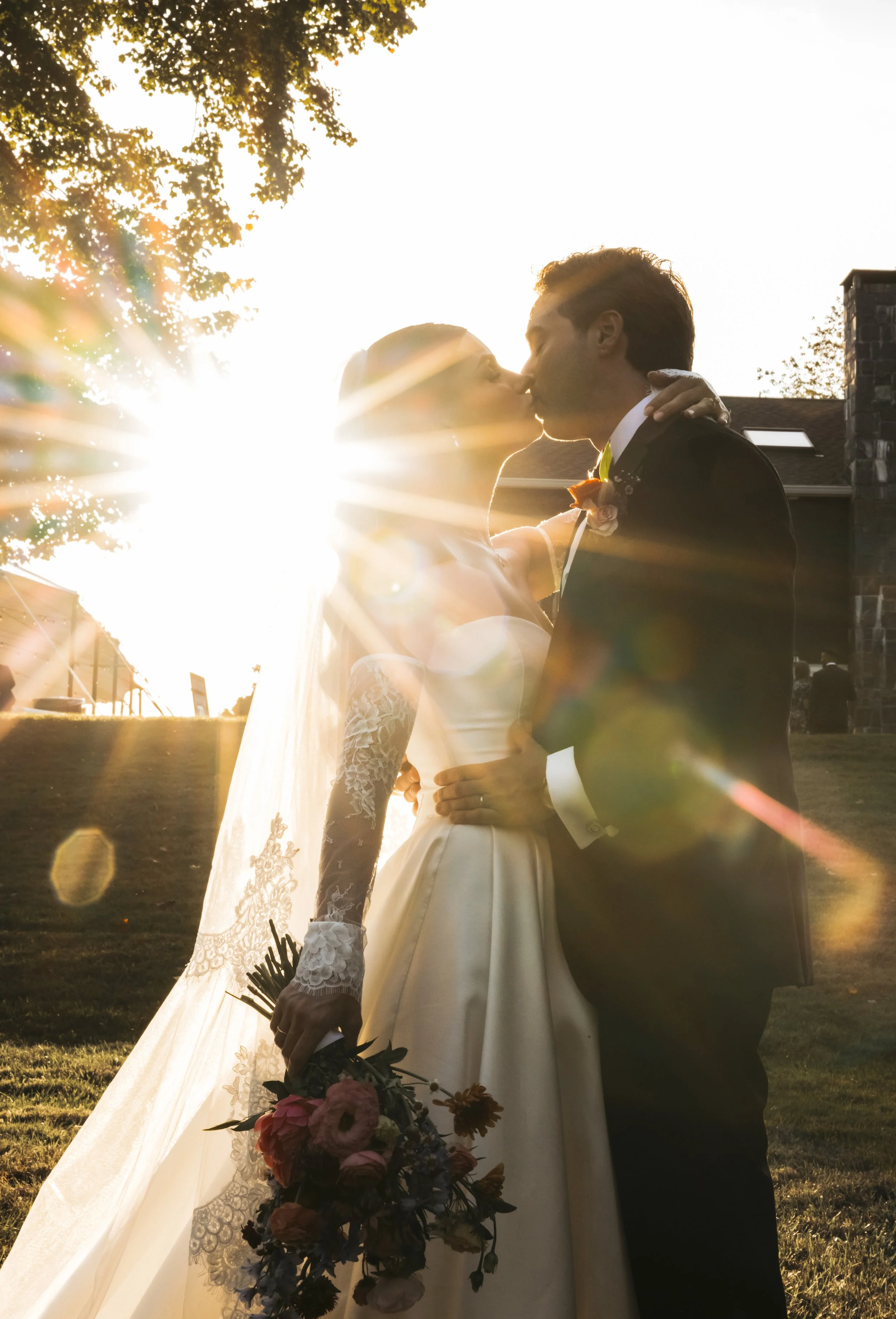 A bride and groom share a kiss outdoors during sunset, with the sunlight creating a bright glow around them and lens flare effects.