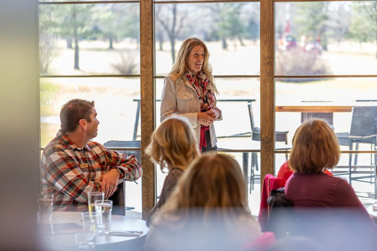 A woman gives a talk to a group at a luncheon with windows behind her showing a golf course.