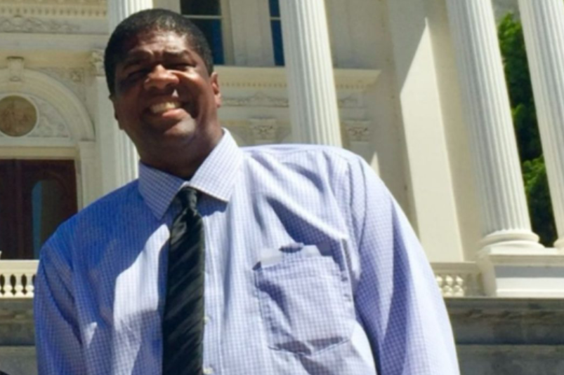 A person in a blue checkered shirt and tie stands outside a large white building with columns.