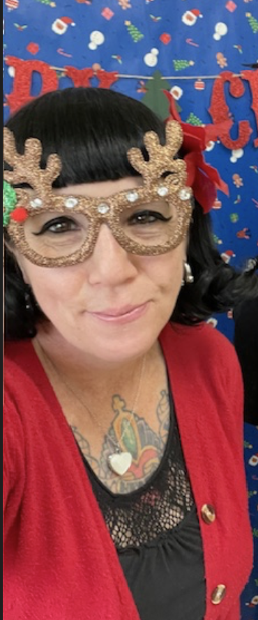 Woman sitting on a chair, wearing a polka dot shirt, black cardigan, and black pants, with hands resting under chin, surrounded by others in a group setting.