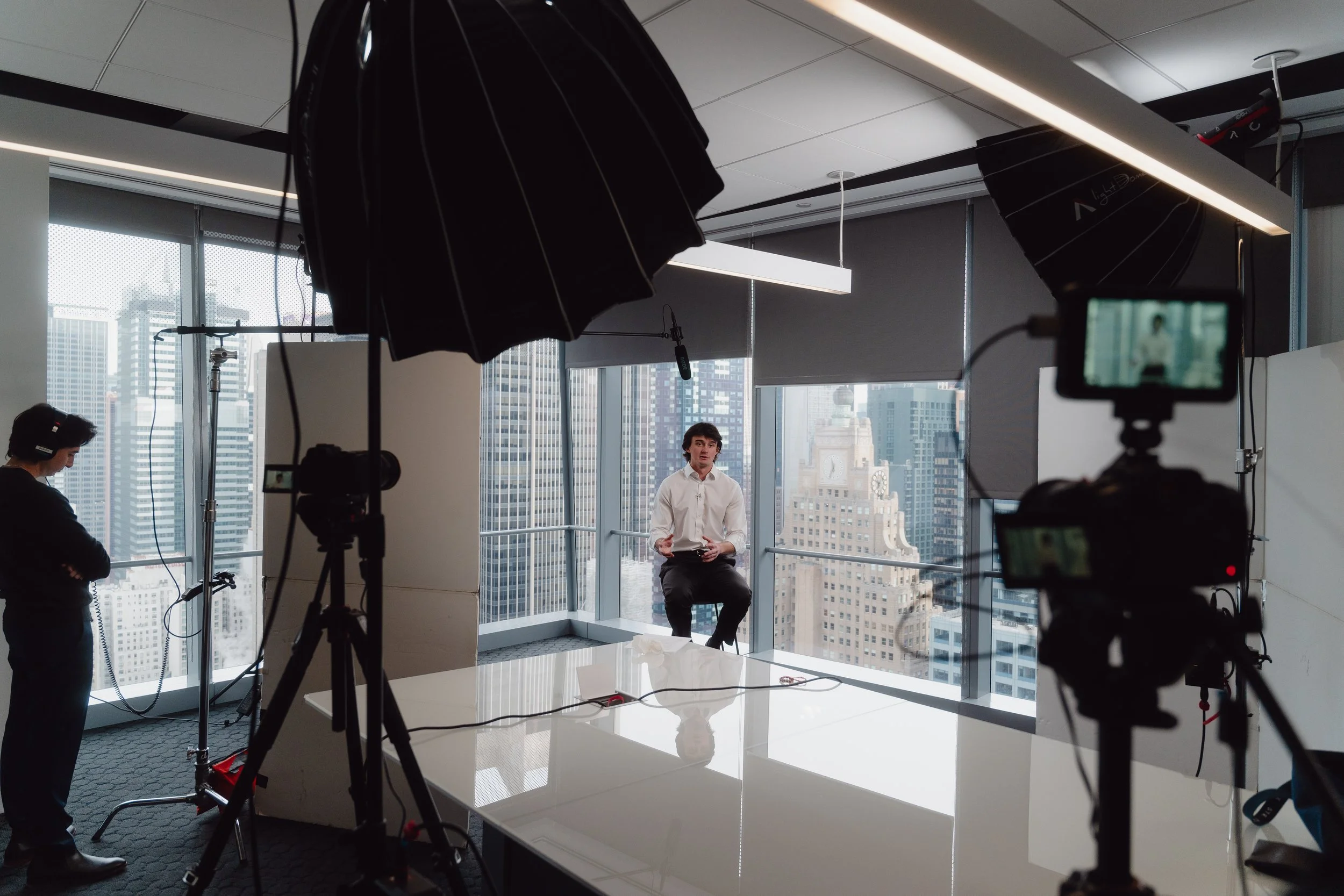 A video interview setup in a high-rise office with large windows and city view, featuring a man in a white shirt sitting on a stool, video cameras, lights, and crew members preparing.
