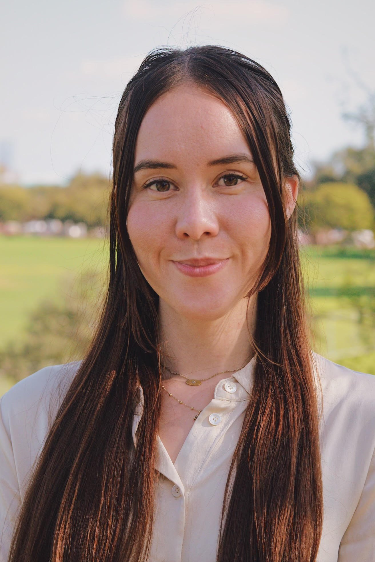 Close-up of a young woman with long brown hair, wearing a white blouse and gold jewelry, standing outdoors with a blurred green park background.