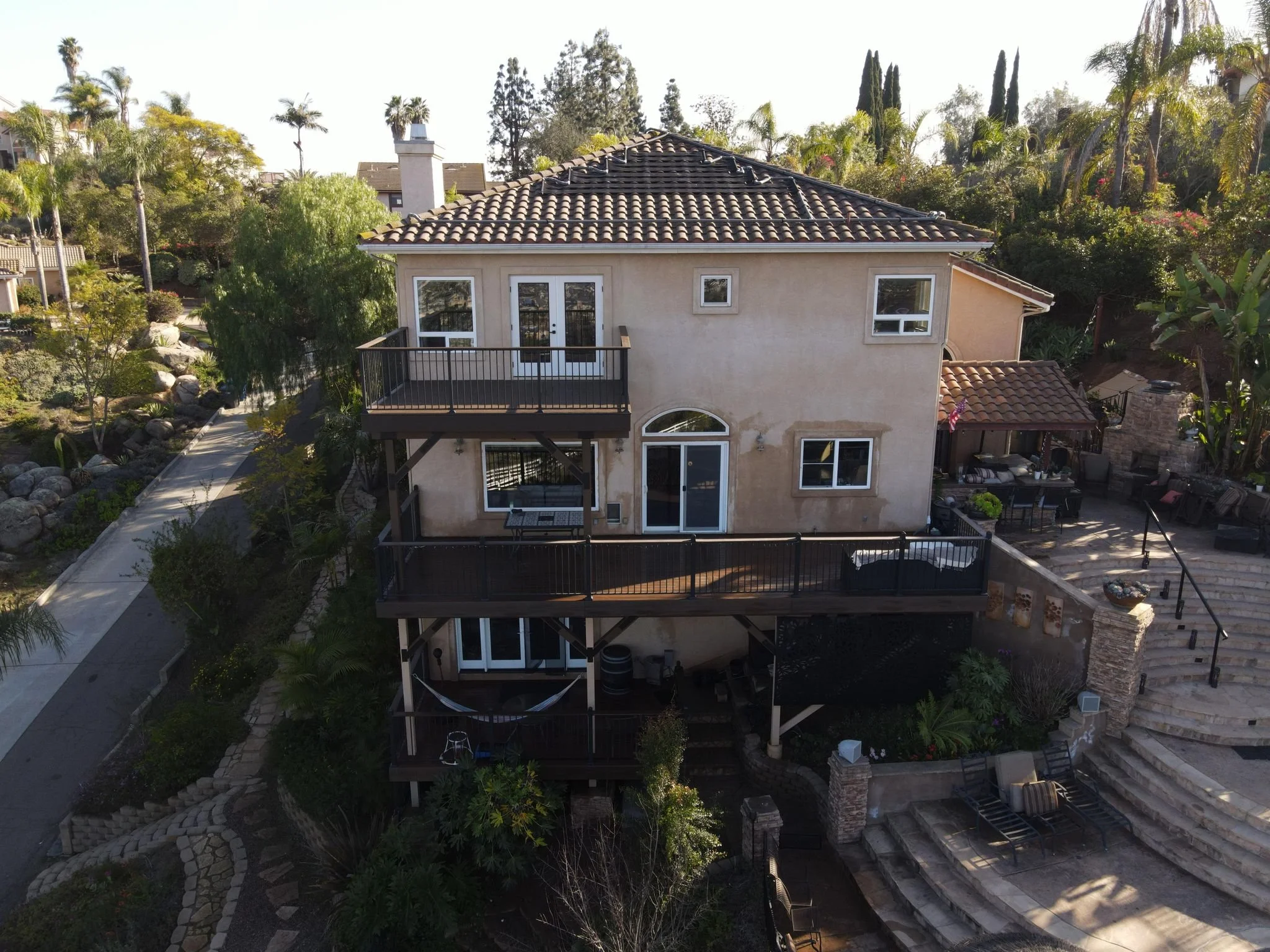 A three-story house with beige walls, multiple windows, and a tiled roof, featuring two balconies with black railings. The house is surrounded by lush landscaping, including palm trees, bushes, and flowers. There is a large staircase on the right side leading to an outdoor patio with seating and a fireplace.