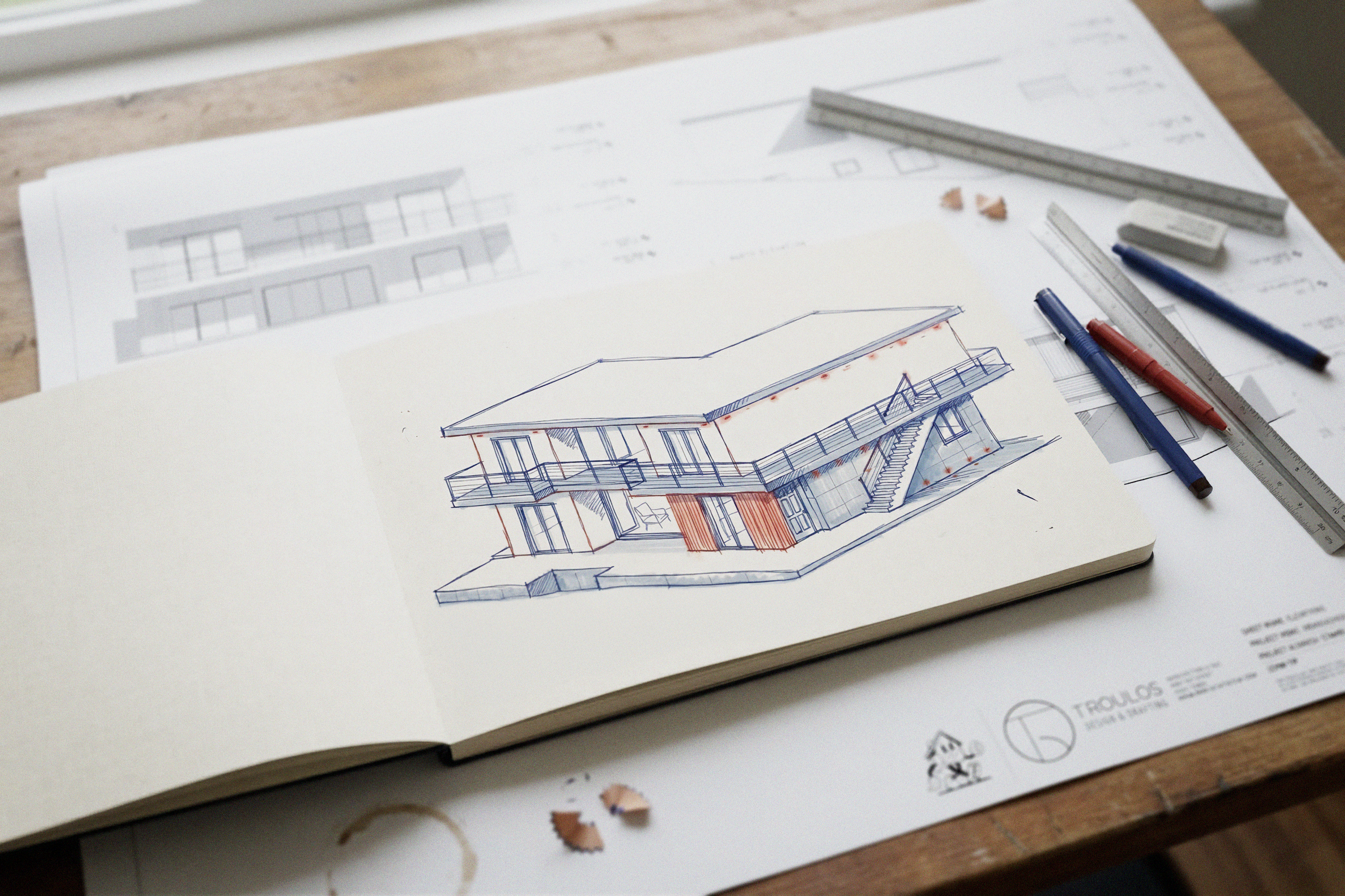 An architectural sketch of a modern house with multiple levels, balconies, and exterior stairs on top of construction plans, on a wooden table with drawing tools.