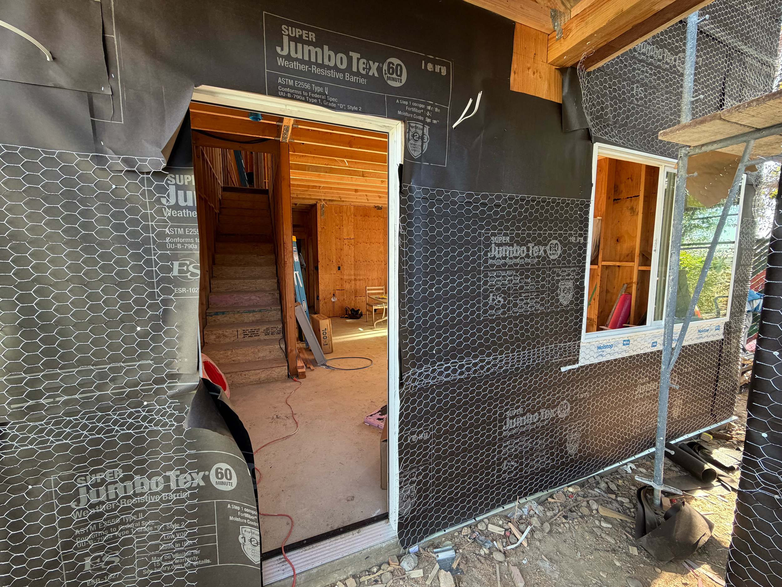 Construction site of a house wall with black weather-resistant barrier, a doorway, and a window frame, including wooden beams and scaffolding.