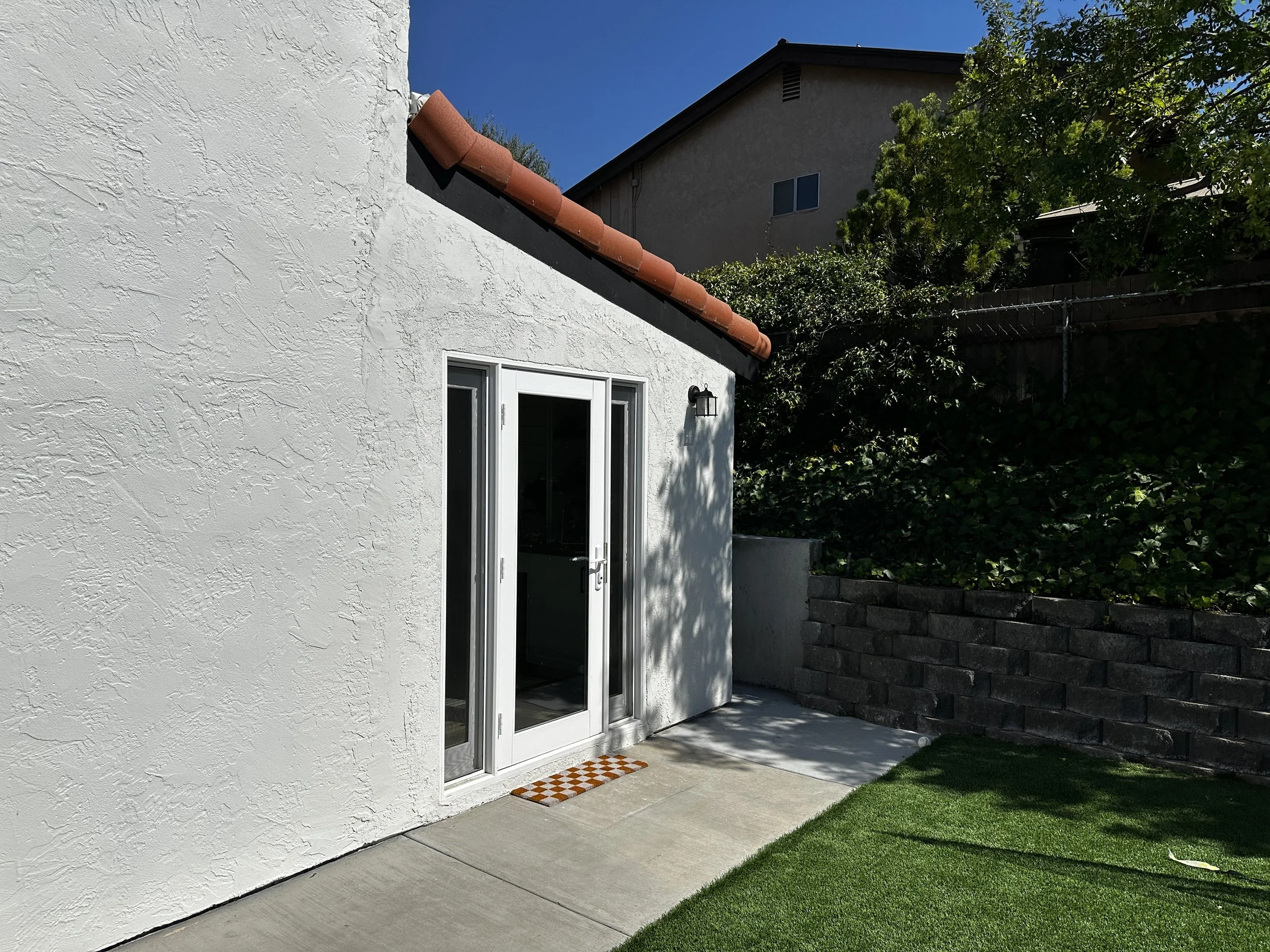 Backyard with white stucco house, sliding glass door, small orange and white checkered doormat, green grass, tree, brick wall, and neighboring houses.