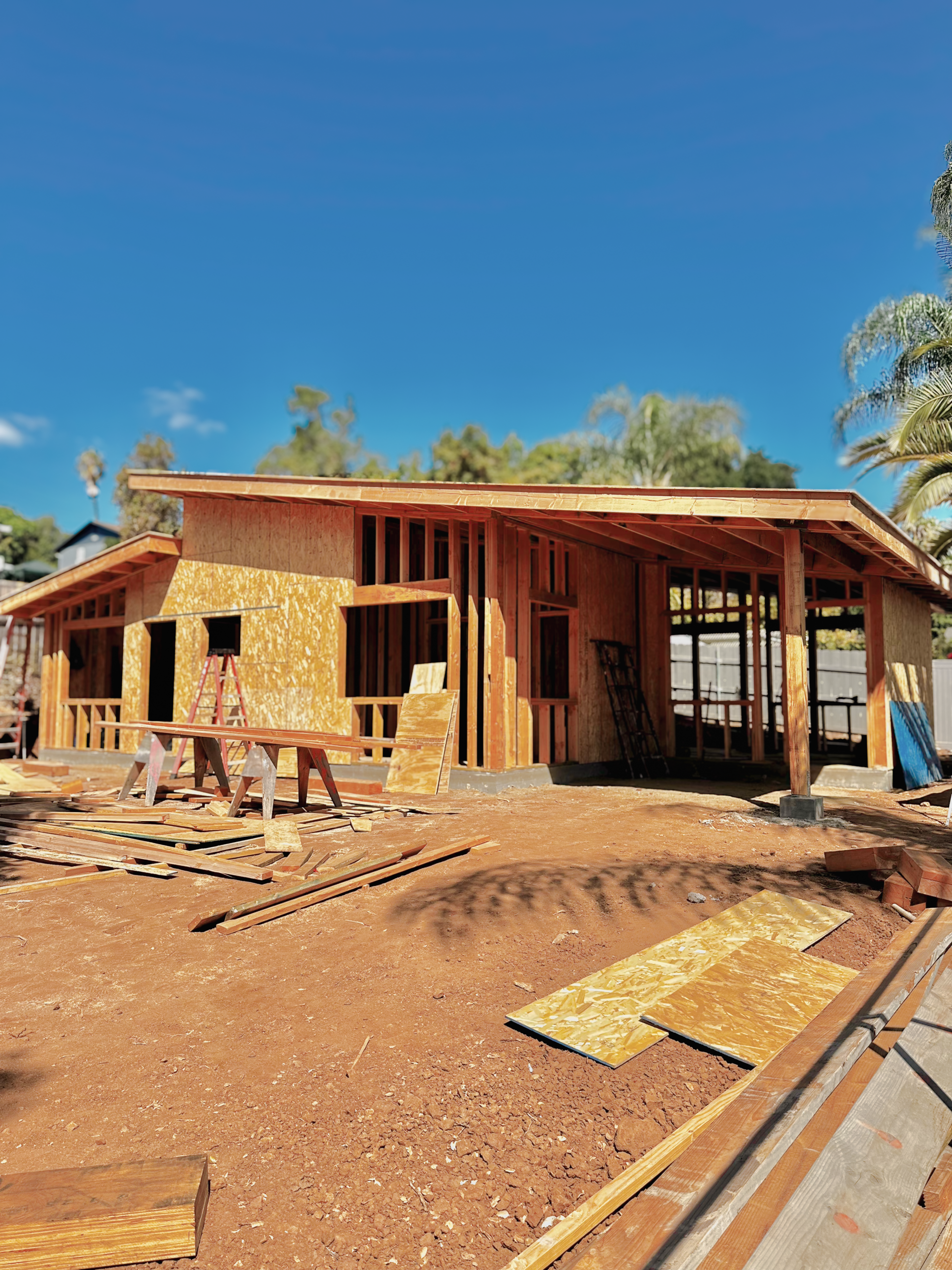 The image depicts a house under construction with wooden framing, construction materials, and tools scattered around on a dirt ground, under a clear blue sky.