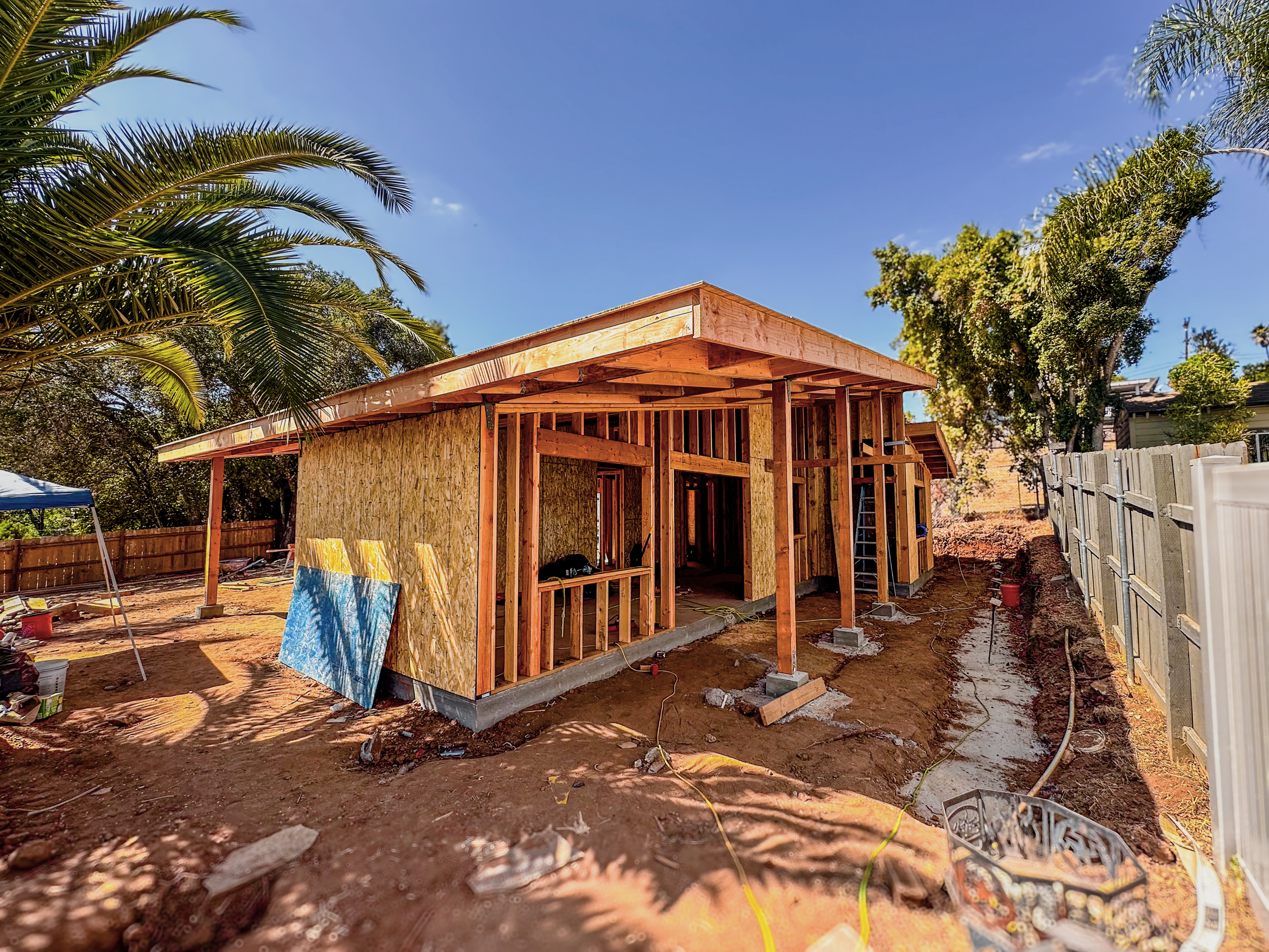 A house under construction with wooden framing, plywood wall sheathing, and a raised concrete foundation, situated outdoors on a dirt lot with trees and a blue sky in the background.