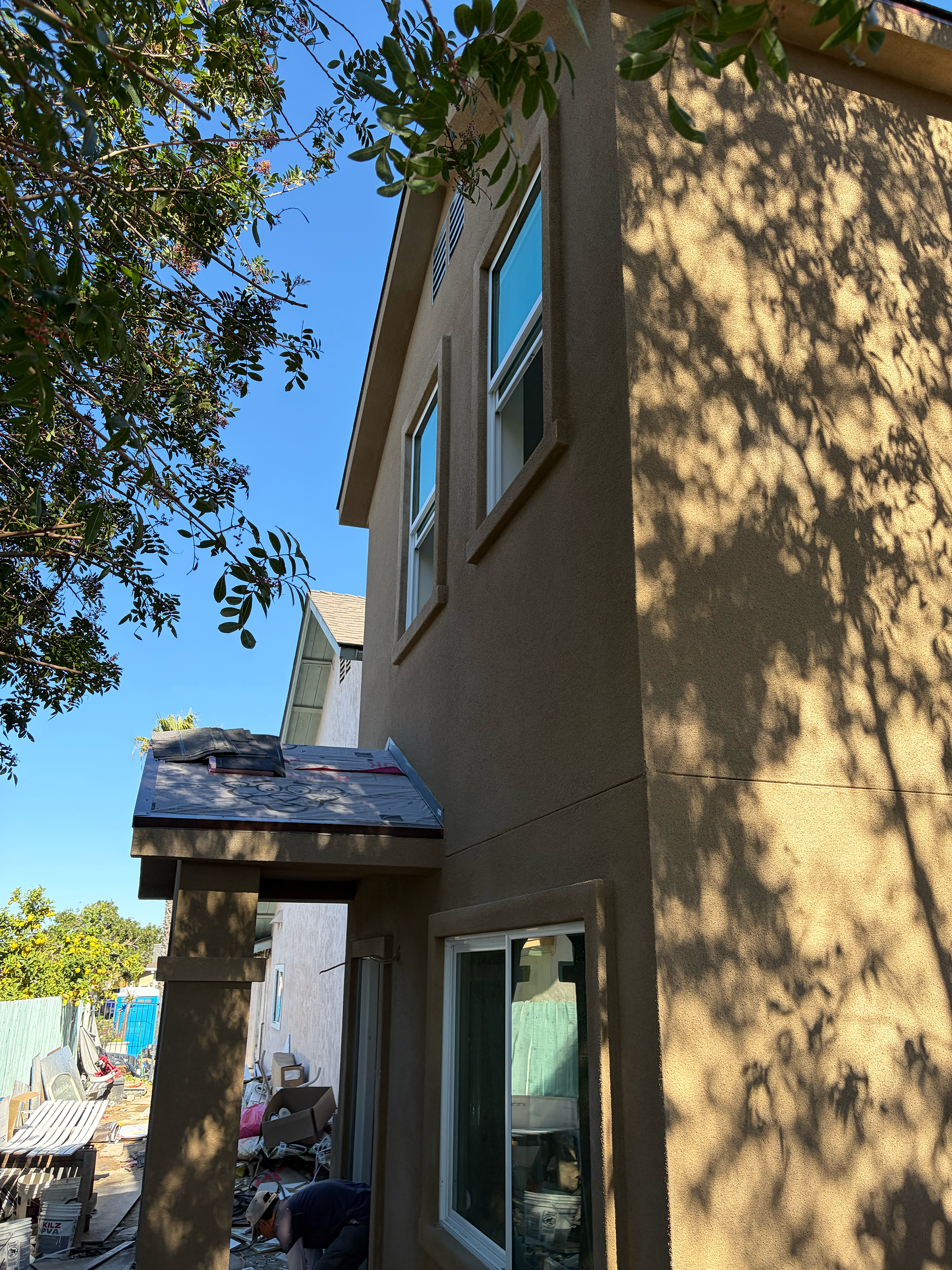 Side view of a multi-story house with two windows on each level, a small overhang above the entrance, and construction materials and debris on the ground, with shadowed trees casting shadows on the wall under a clear blue sky.