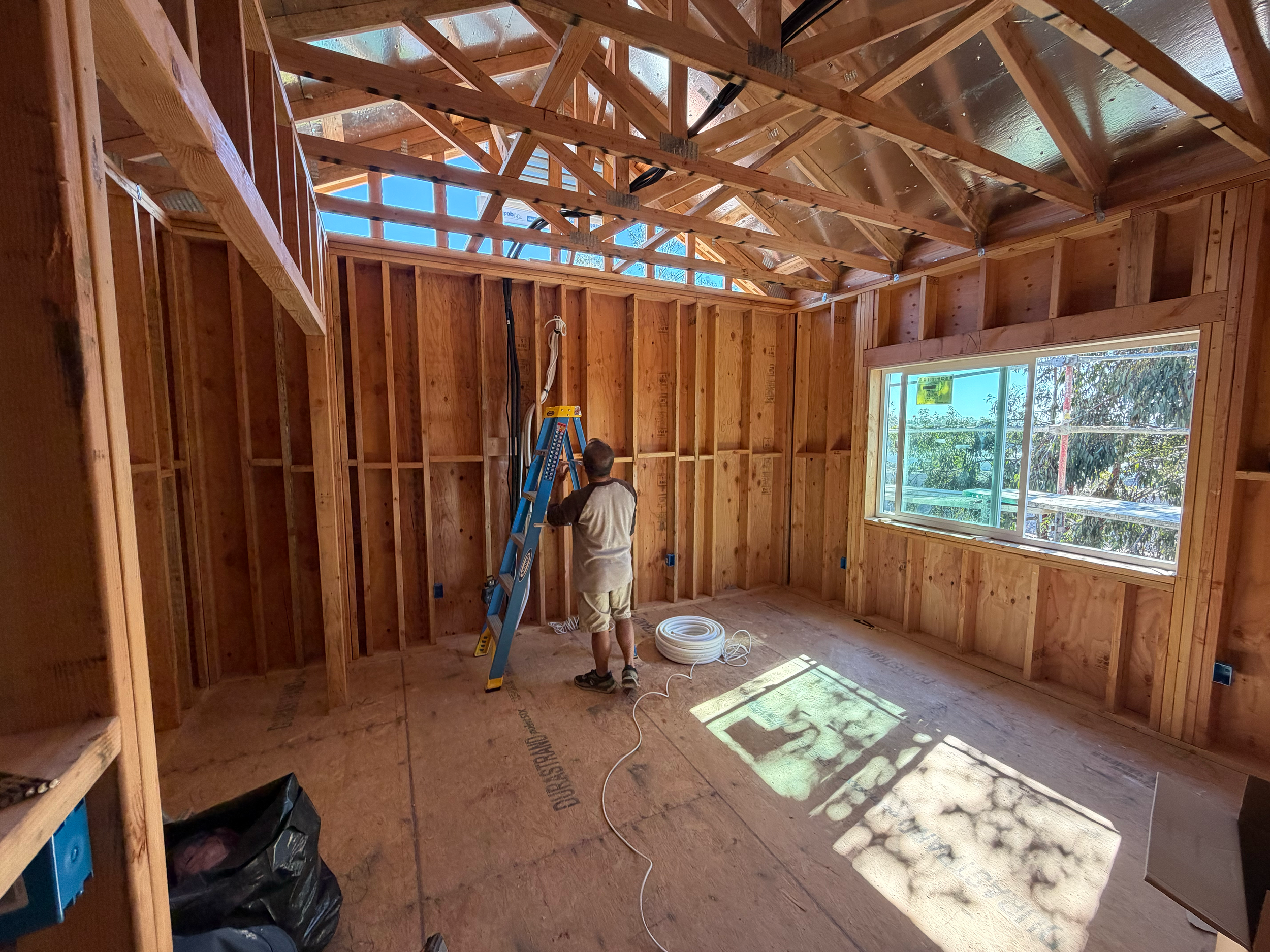 A person working on electrical wiring in a house under construction, with exposed wooden framing, a window, and sunlight casting shadows on the floor.