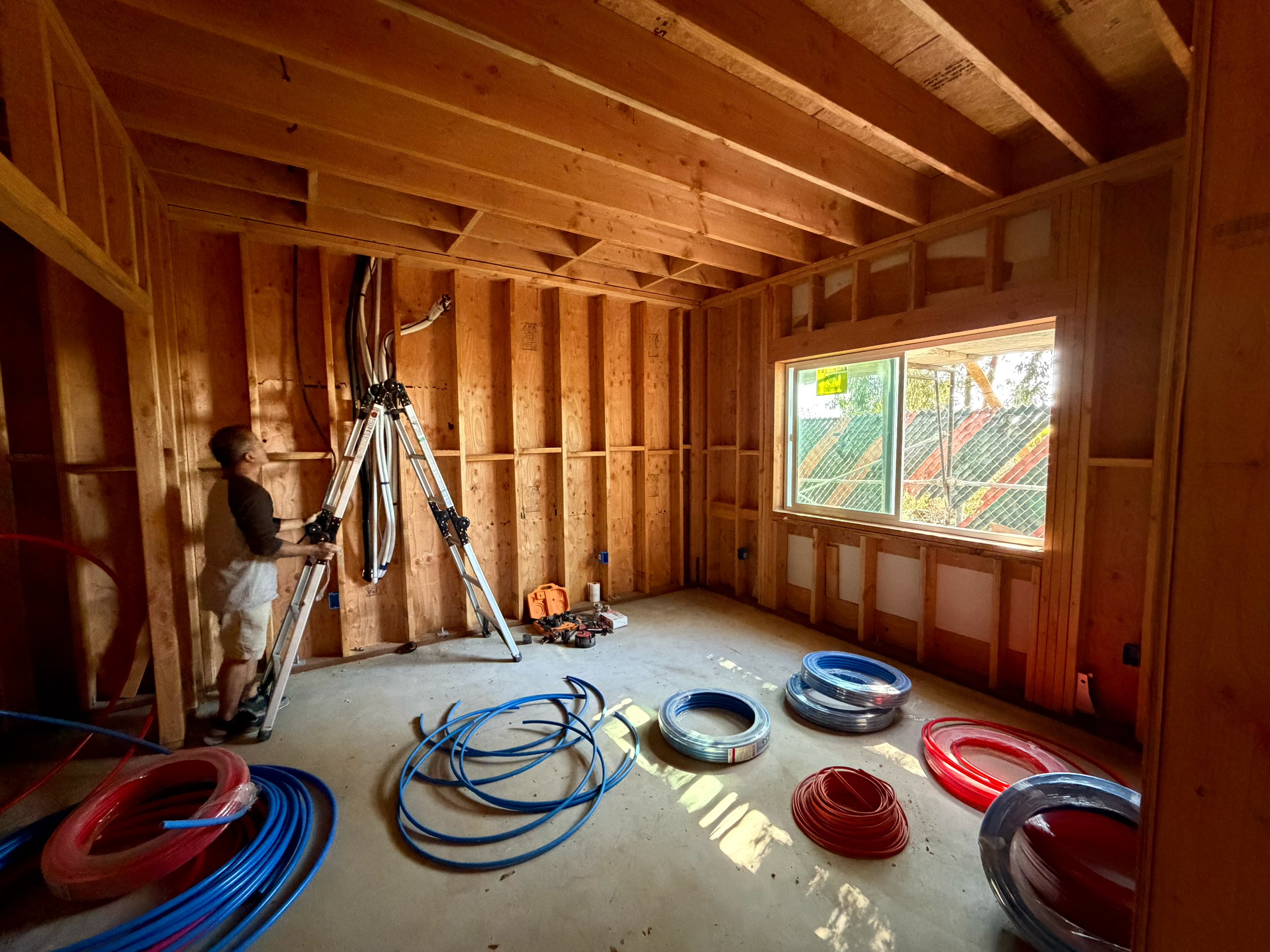 Interior of a house under construction with walls and ceiling framing visible. A person is working on electrical wiring with a ladder. Coiled wires in red and blue are on the floor near a window.