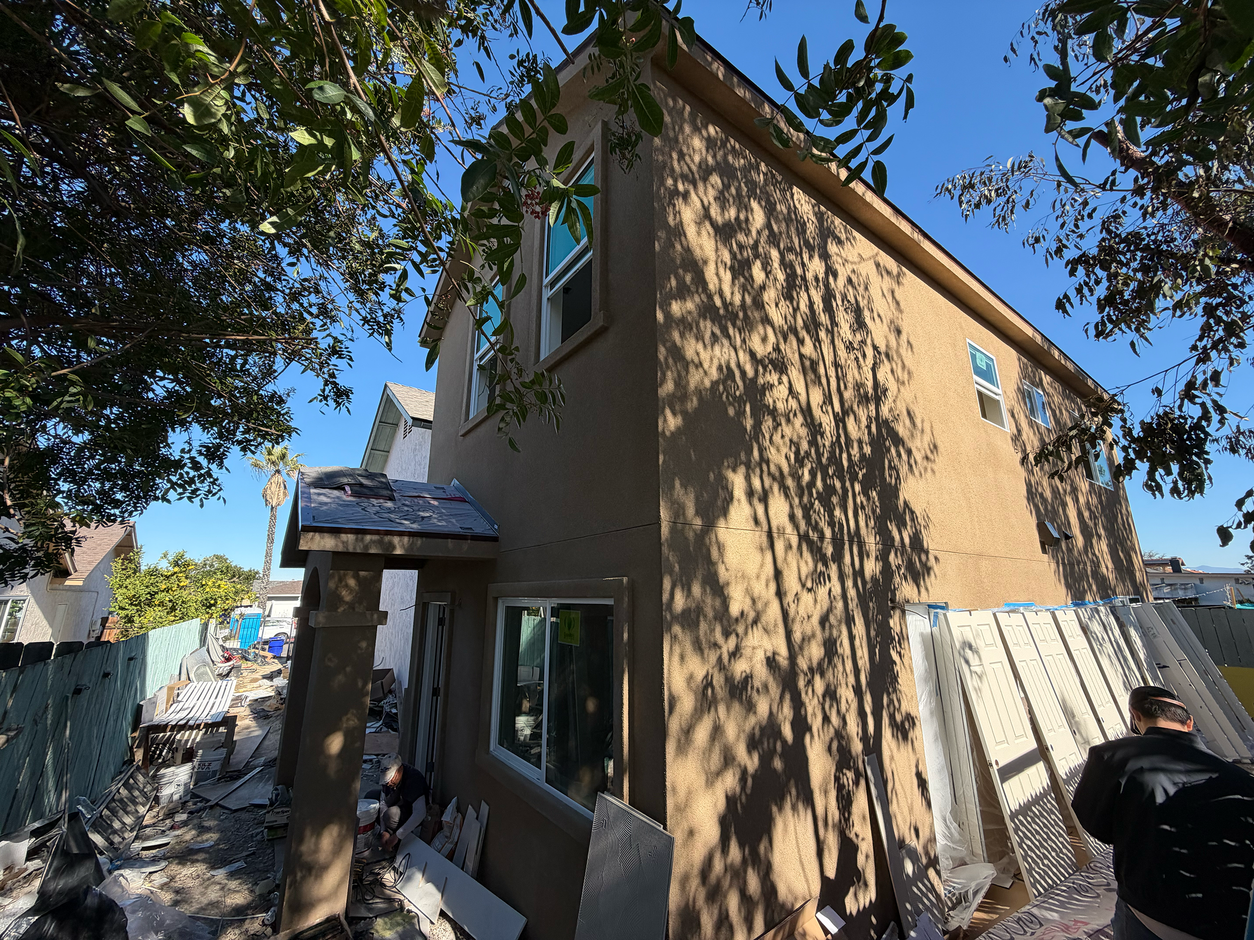 Construction site of a two-story beige house with workers installing exterior siding, surrounded by trees and construction materials.