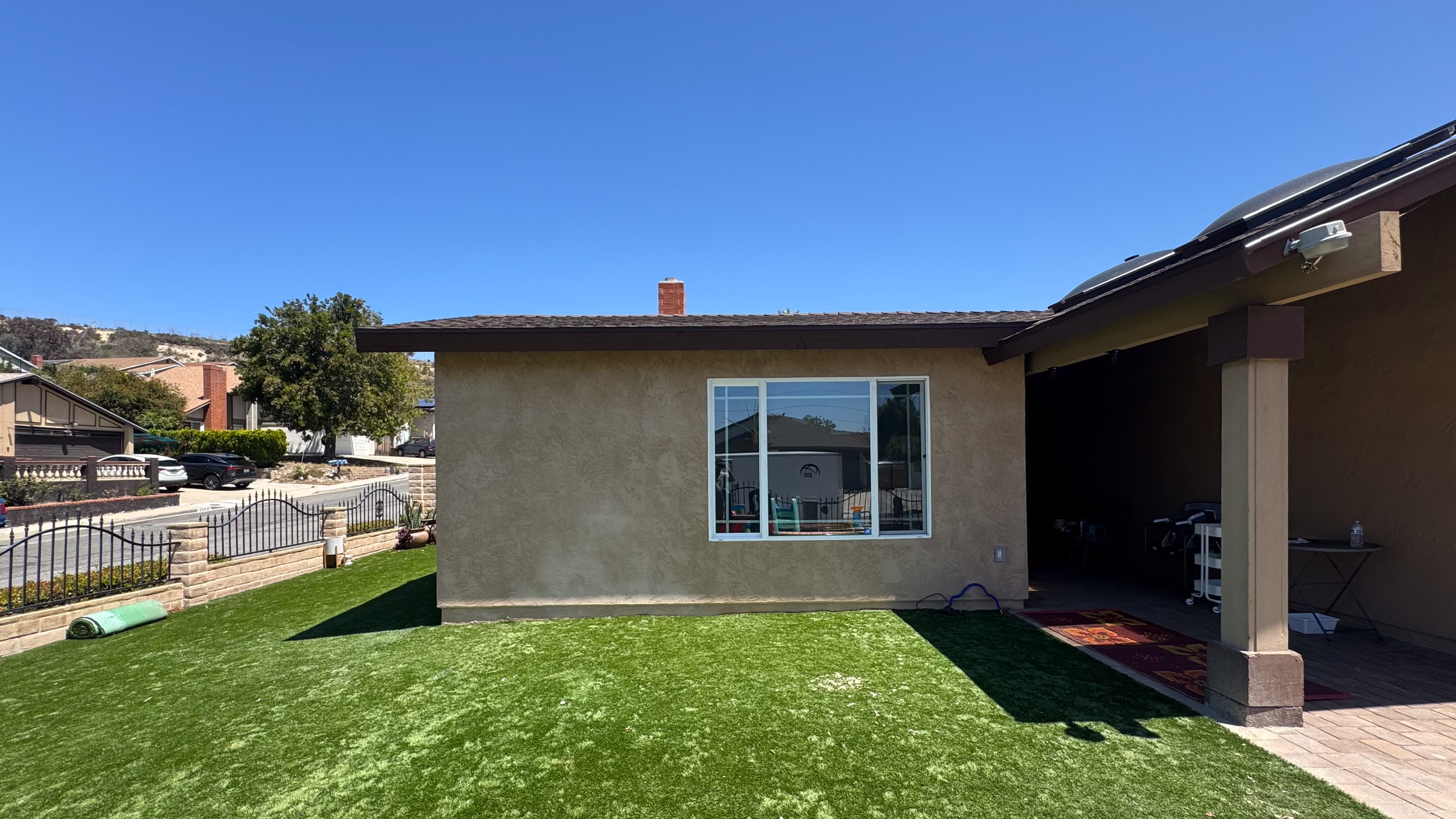 Backyard with a well-maintained green lawn, beige house wall with a window, and a patio covered area with chairs and a small table, under a clear blue sky.