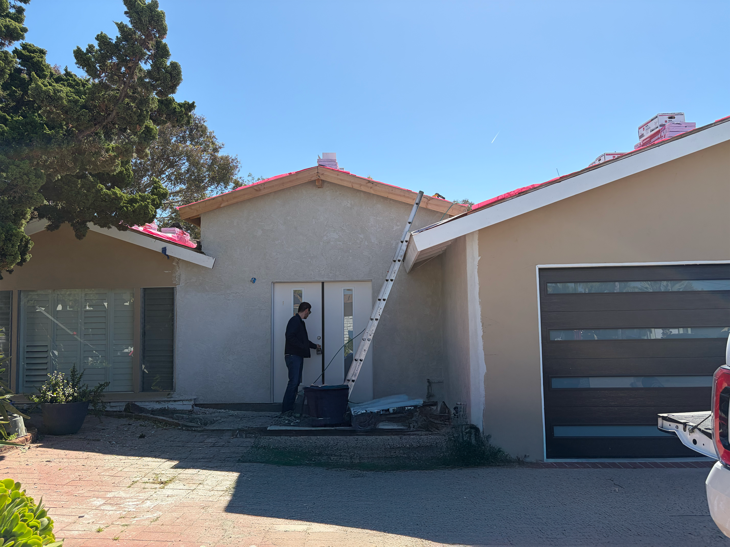 A person standing outside a house during construction or renovation, with a ladder leaning against the roof, and construction materials on the roof. The house has a beige exterior, large windows, and a black garage door, with a car parked on the driveway.