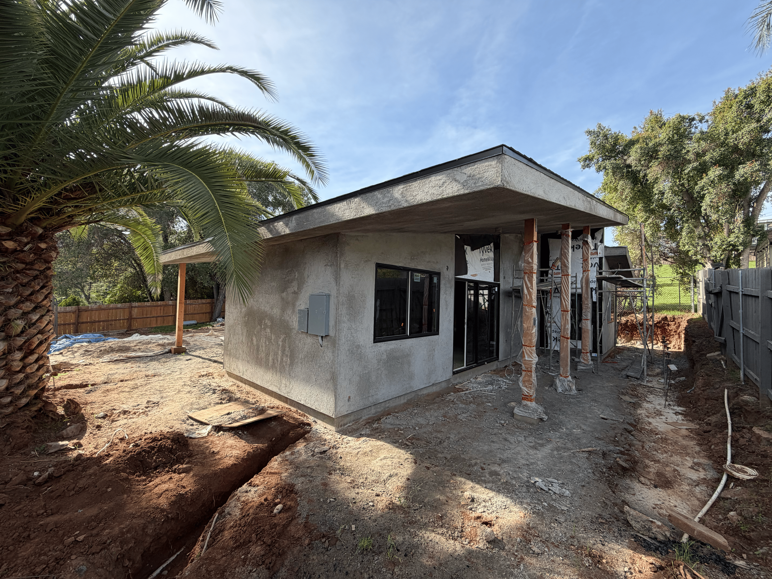 Construction site of a small house with gray walls, black window frames, and front door. Two temporary wooden supports hold up the front roof section. Scaffolding is present beside the house. The yard has exposed dirt, a large palm tree on the left, and a wooden fence in the background. Clear blue sky is overhead.