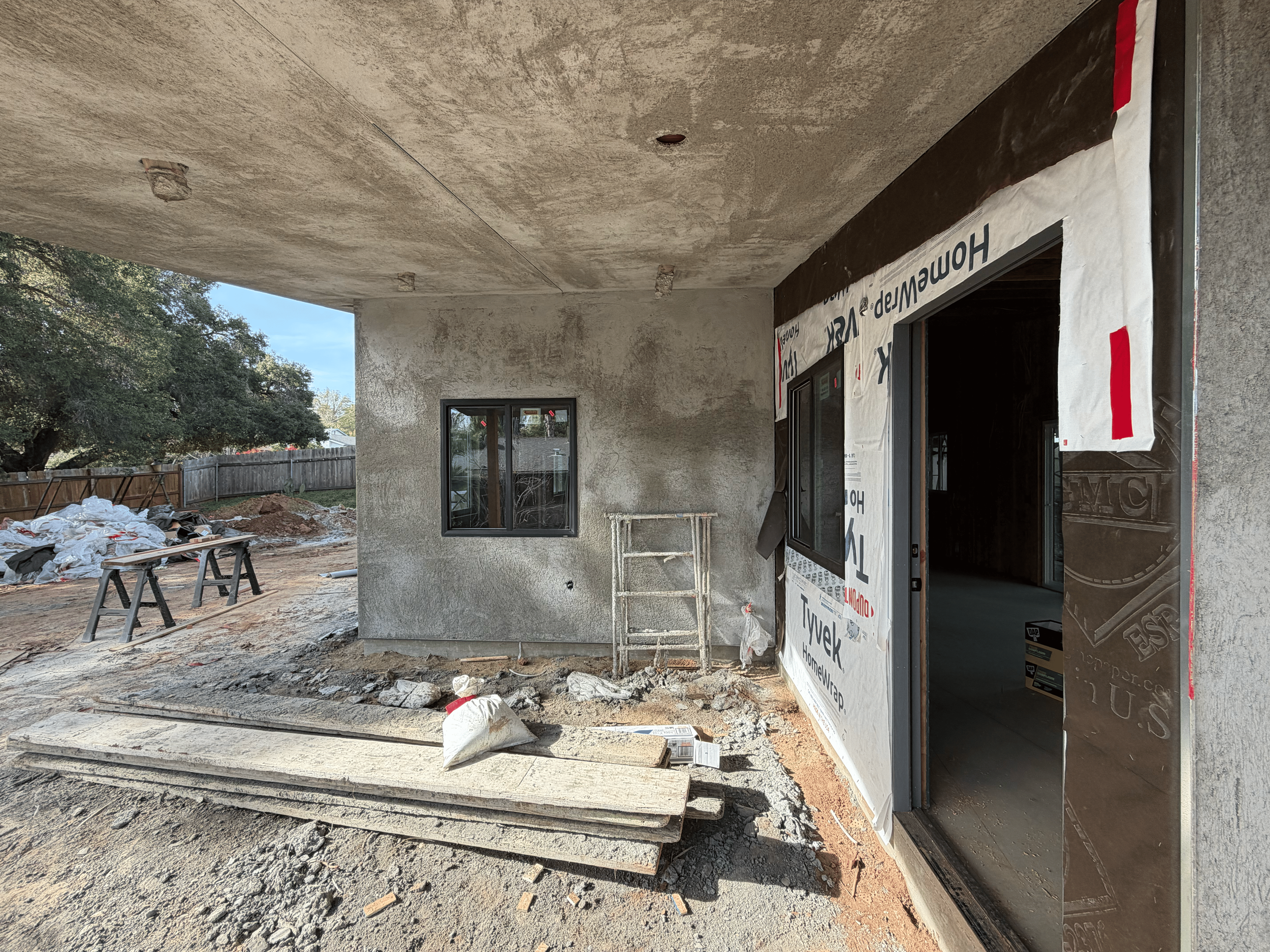 Exterior of a house under construction with a doorway, window, building materials, and tools visible. The house is partially finished with insulation and wall coverings installed.
