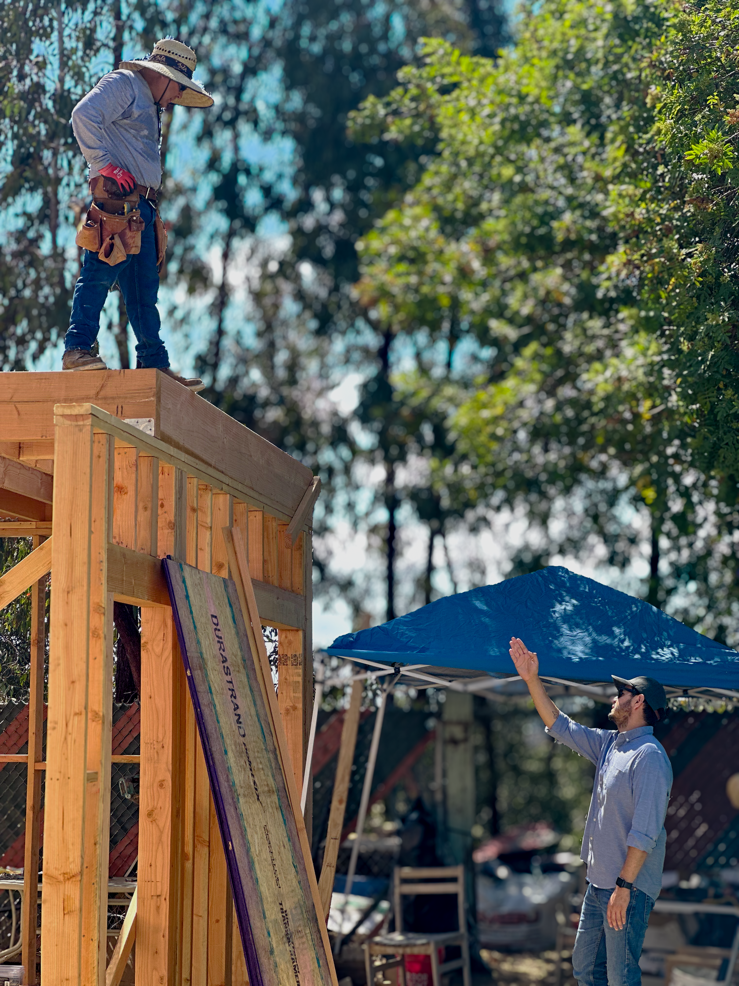 A construction worker wearing a straw hat, gloves, and a tool belt stands on a wooden platform, looking down at a man waving from below at a construction site with a blue canopy and trees in the background.