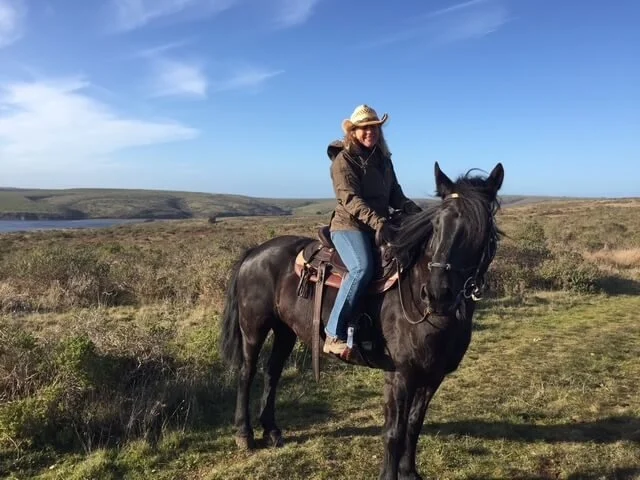 A woman riding a black horse on a grassy plain under a blue sky with some clouds.