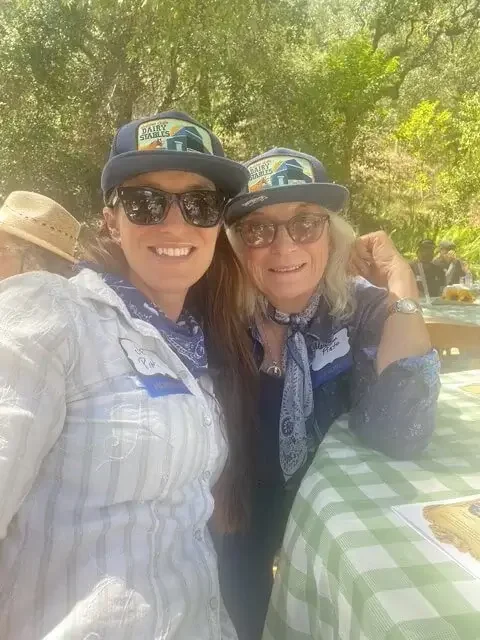 Two women wearing bandanas, sunglasses, and caps with a Texas theme, smiling at an outdoor gathering with a green checkered tablecloth, trees, and other people in the background.