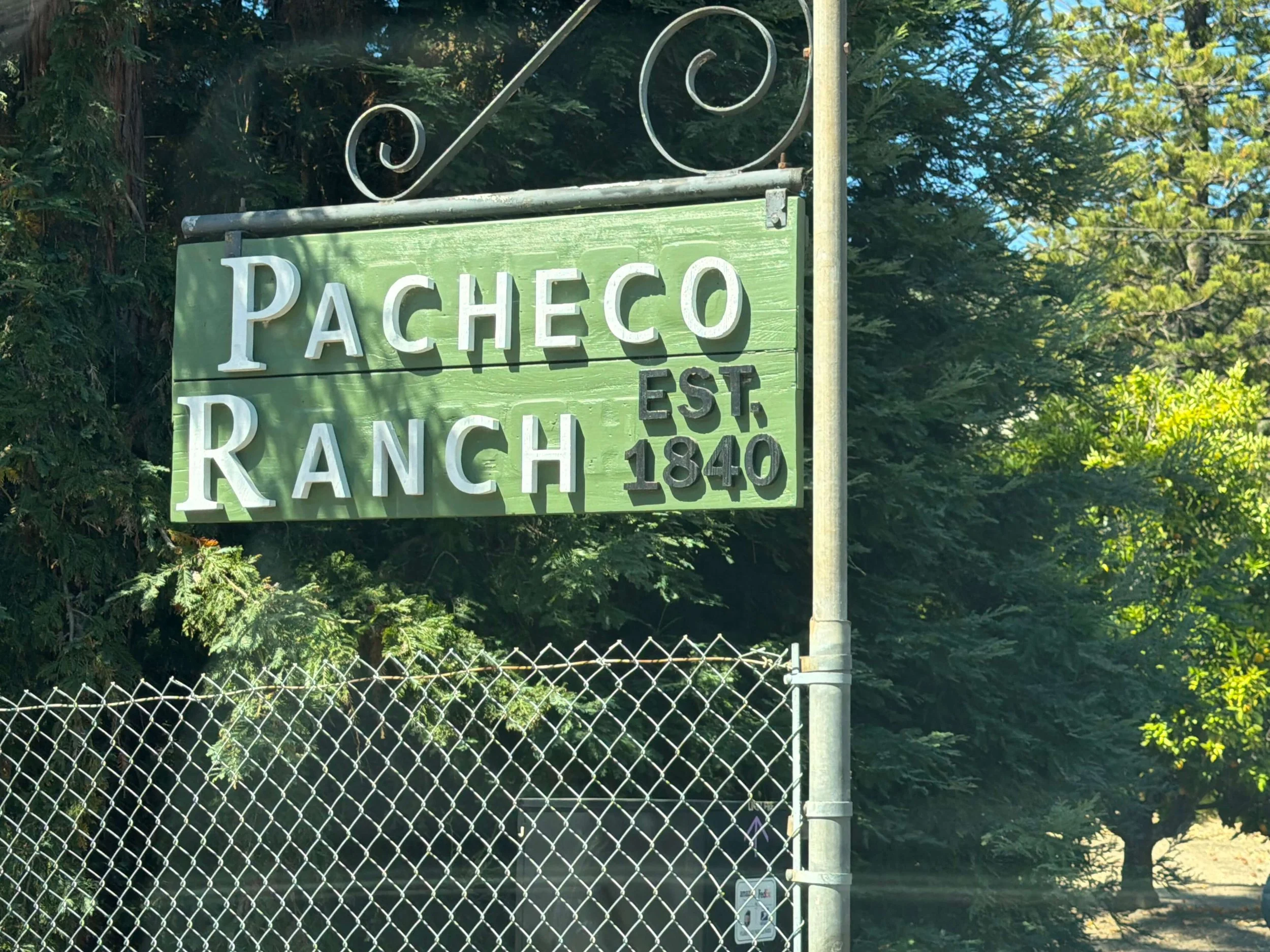 A green street sign with white letters reading 'Pacheco Ranch Est. 1840' attached to a metal pole above a chain-link fence, with trees and blue sky in the background.