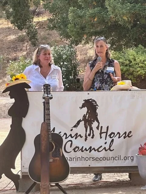 Two women standing behind a table with a sign that reads "Carin Horse Council," outdoors with trees and greenery in the background. One woman is speaking into a microphone, the other looks on. A guitar is leaning against the table, and there are sunflowers on each side of the table.