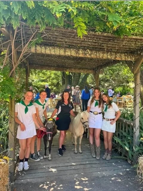 Group of girls and women with white horses under a wooden arbor surrounded by greenery.